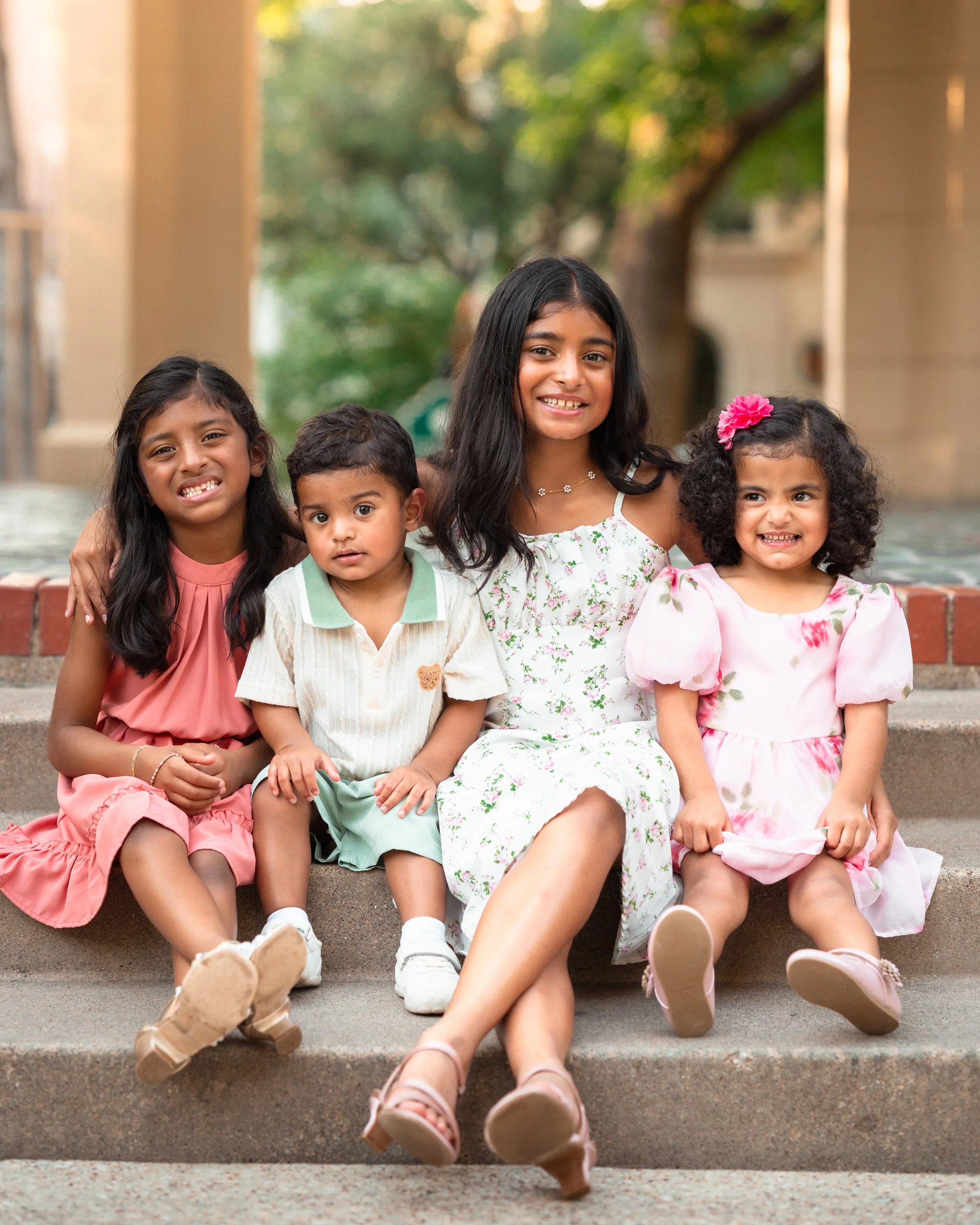 A group of five young girls sitting side by side on steps outdoors, smiling and looking at the camera, with a blurred green tree and building in the background.