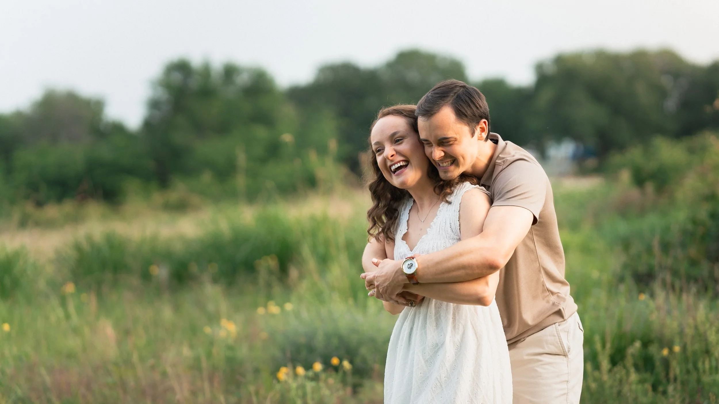 A couple laughing and hugging outdoors in a green field with trees in the background.