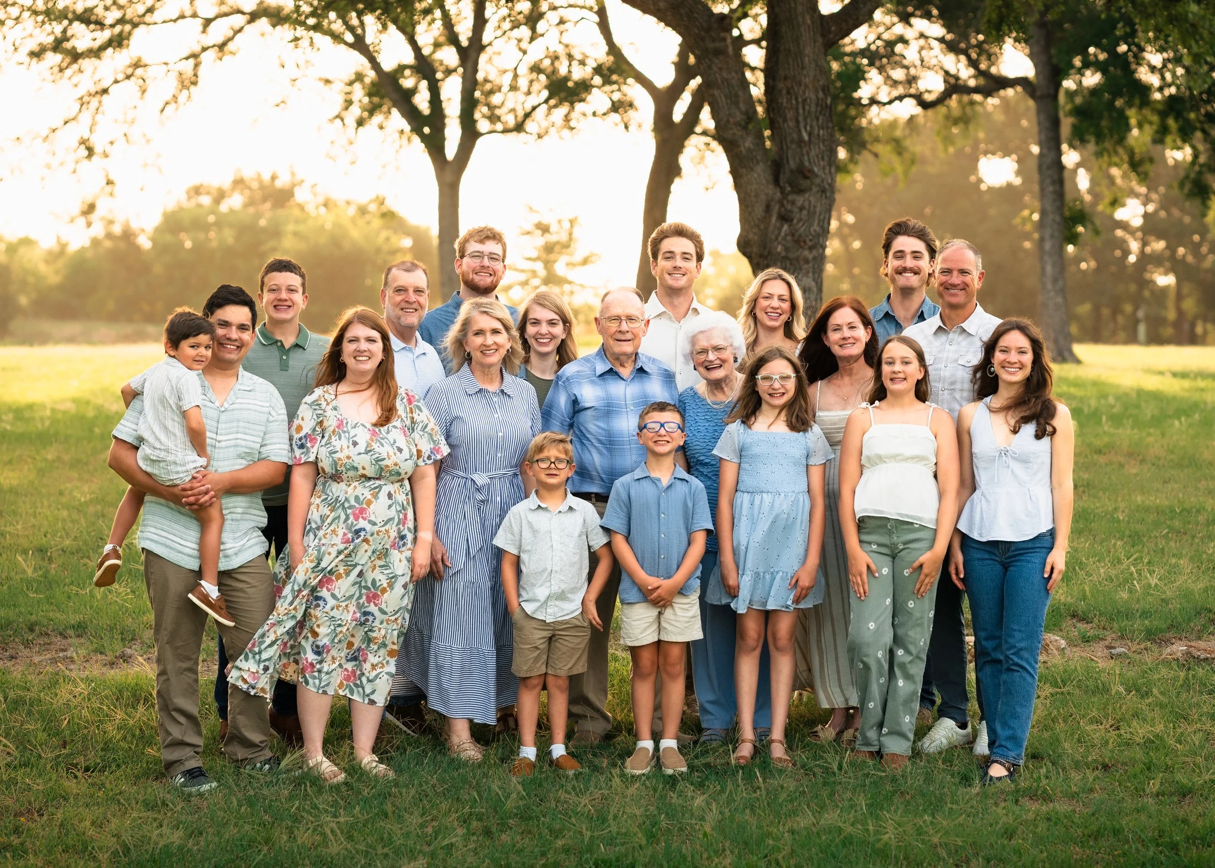 A large, multigenerational family outdoors on a sunny day, standing together on a grassy area with trees in the background, smiling and posing for a group photo.