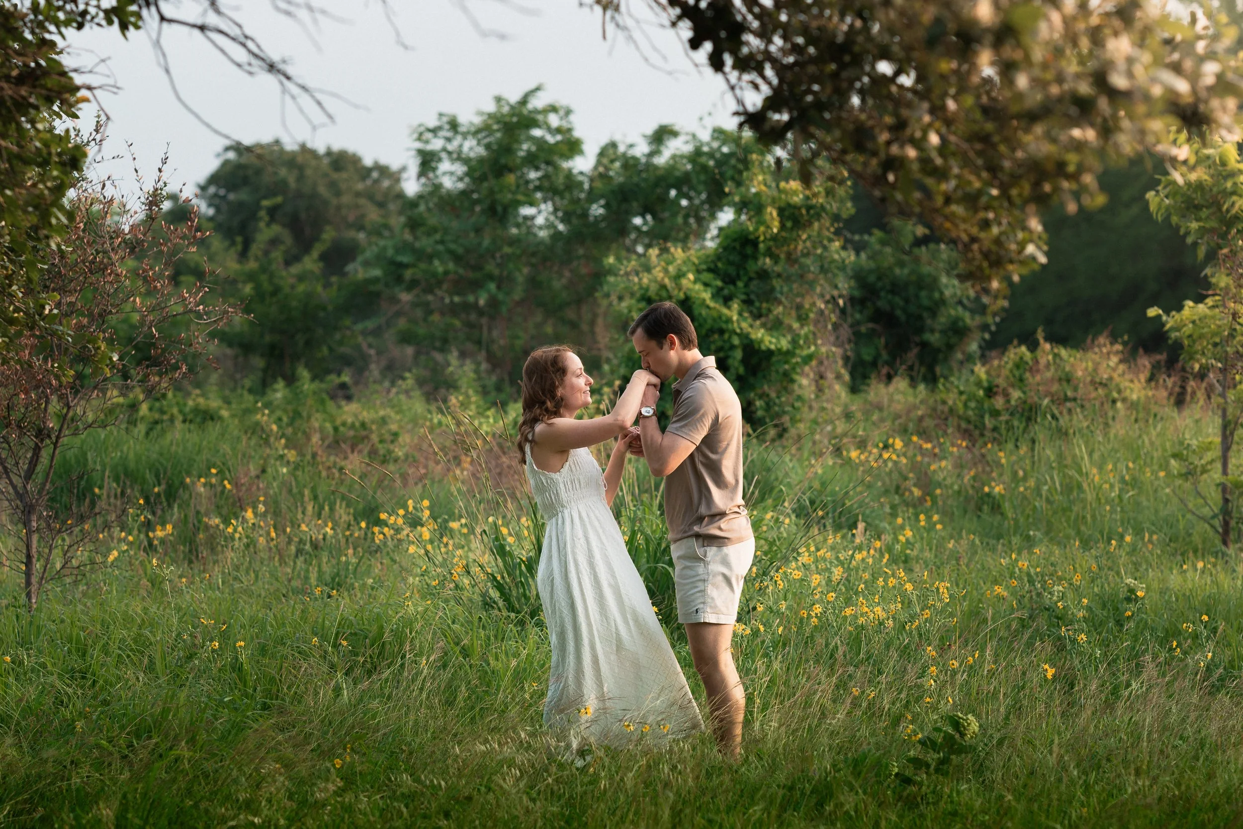 A couple is dancing in a lush green field with trees and wildflowers. The woman is wearing a white dress and the man is dressed casually in a beige shirt and shorts. The man is gently kissing the woman's hand.