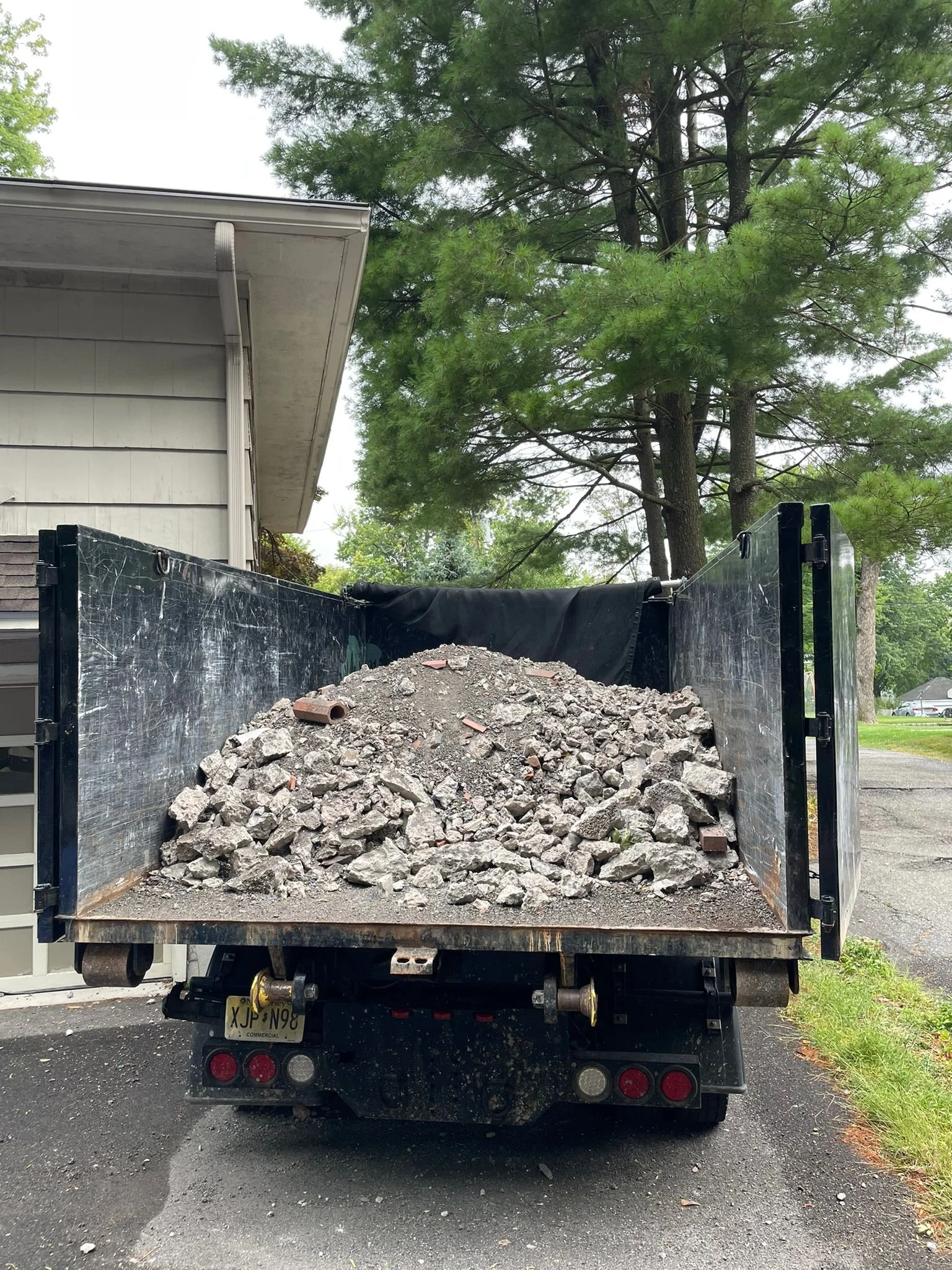 A dump truck loaded with a pile of gravel and rocks parked in front of a house and trees.