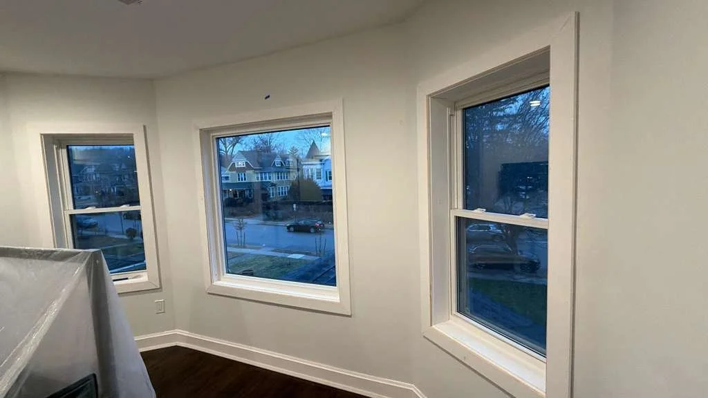 Photo of an interior room showing three large windows with white frames, overlooking a neighborhood street with houses, trees, and parked cars, taken during dusk or early evening.