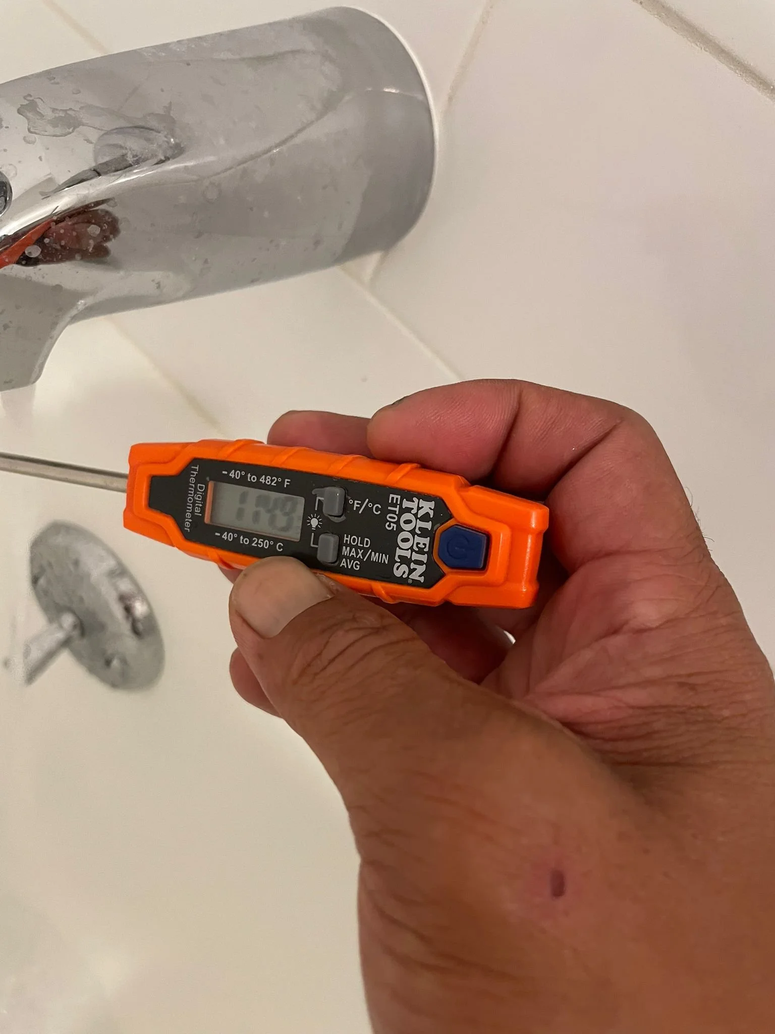 A person holding a digital thermometer near a bathroom shower faucet to measure water temperature.