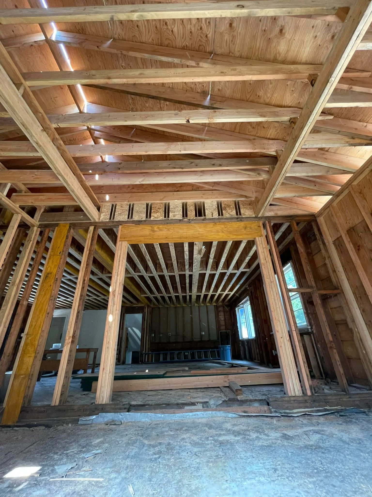 Interior of a house under construction with exposed wooden framing and ceiling, bare floor, and windows in the background.