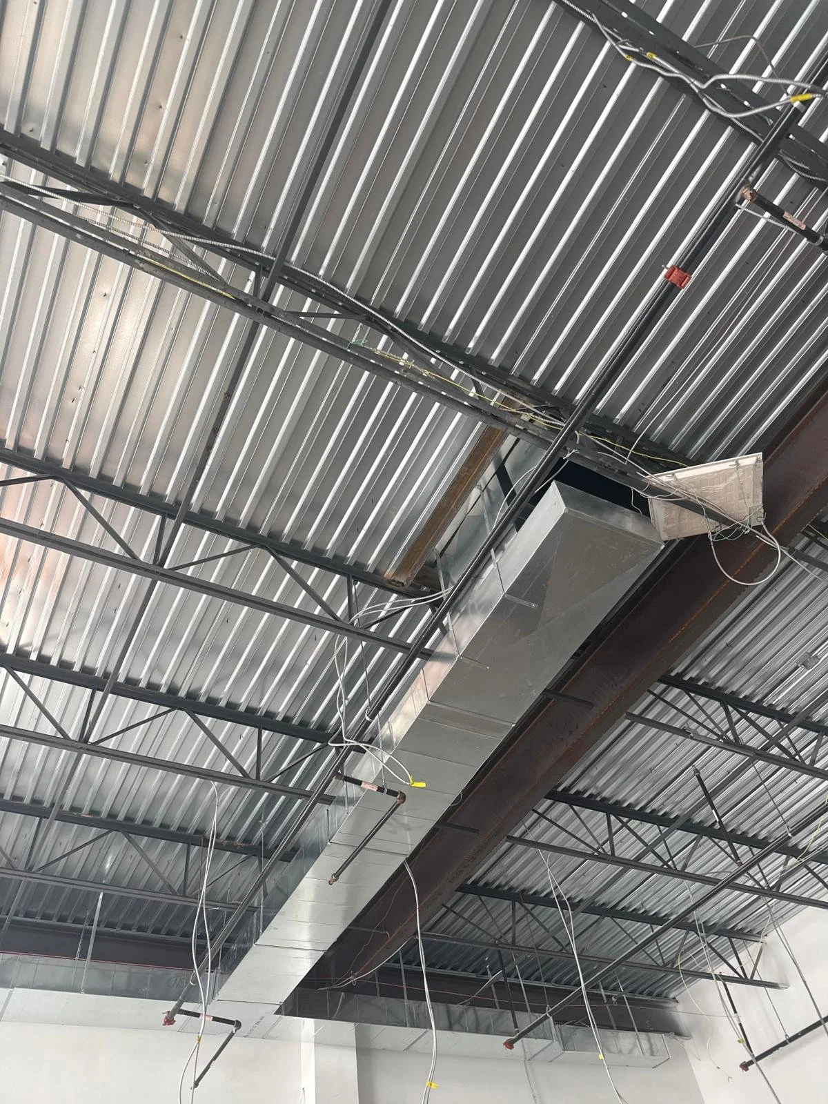 Ceiling of an industrial or commercial building with exposed metal ductwork, piping, and electrical wiring.