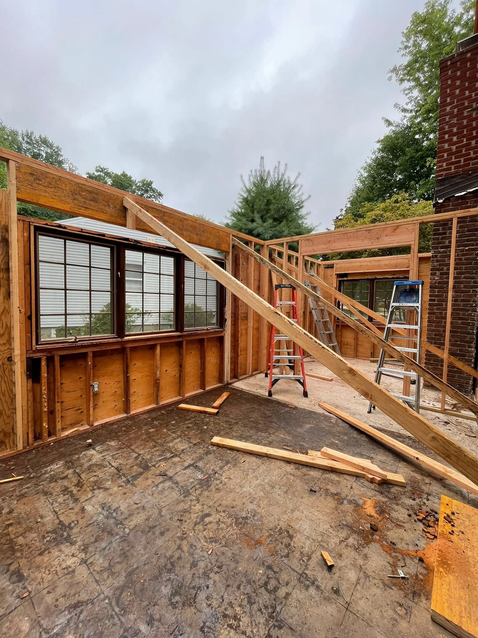 A house under construction with wooden framing, ladders, and a large window on a cloudy day.