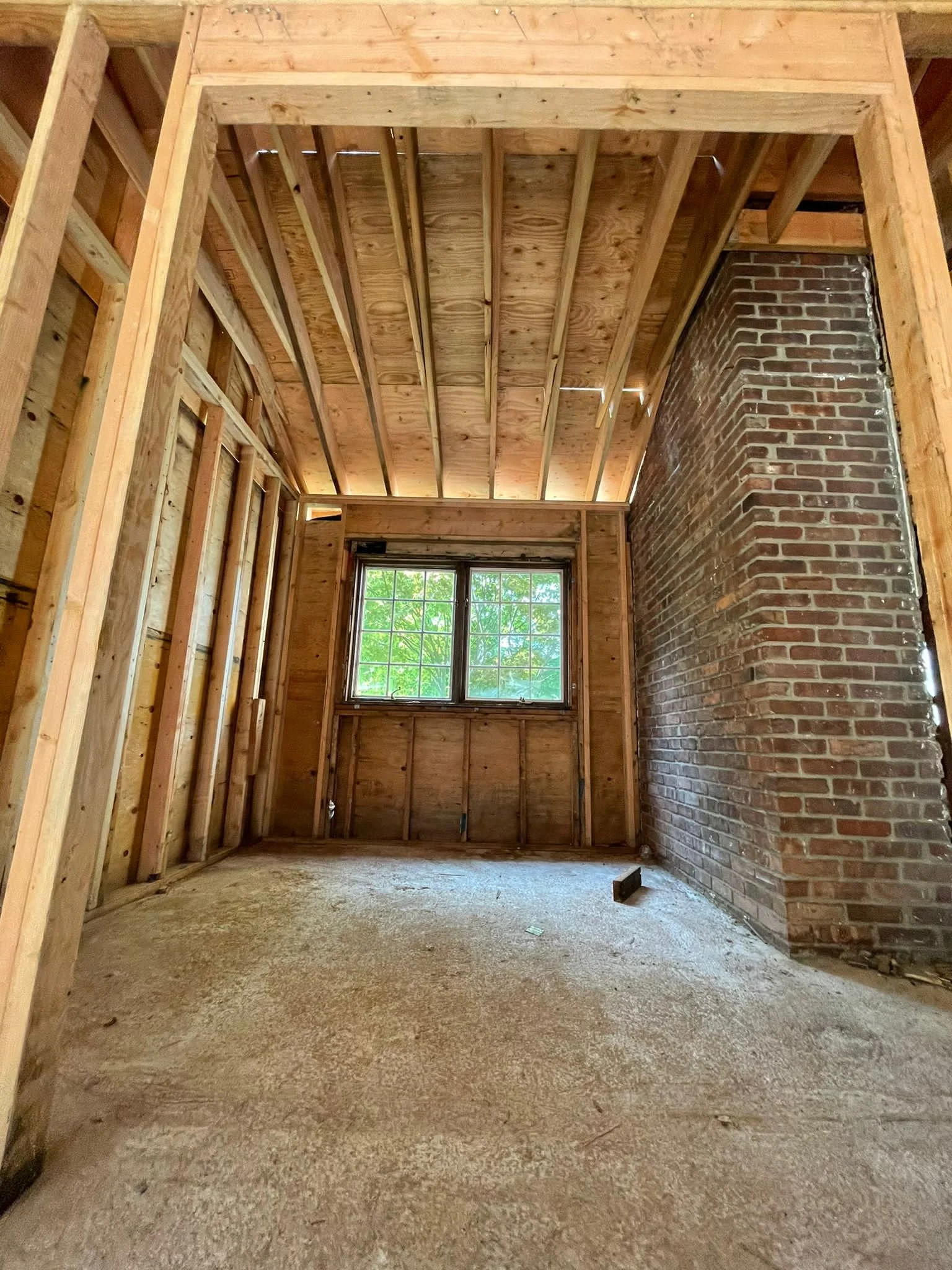 Interior of a room under construction with exposed wooden framing, a brick chimney on the right, a window at the far end, and a roughly finished floor.