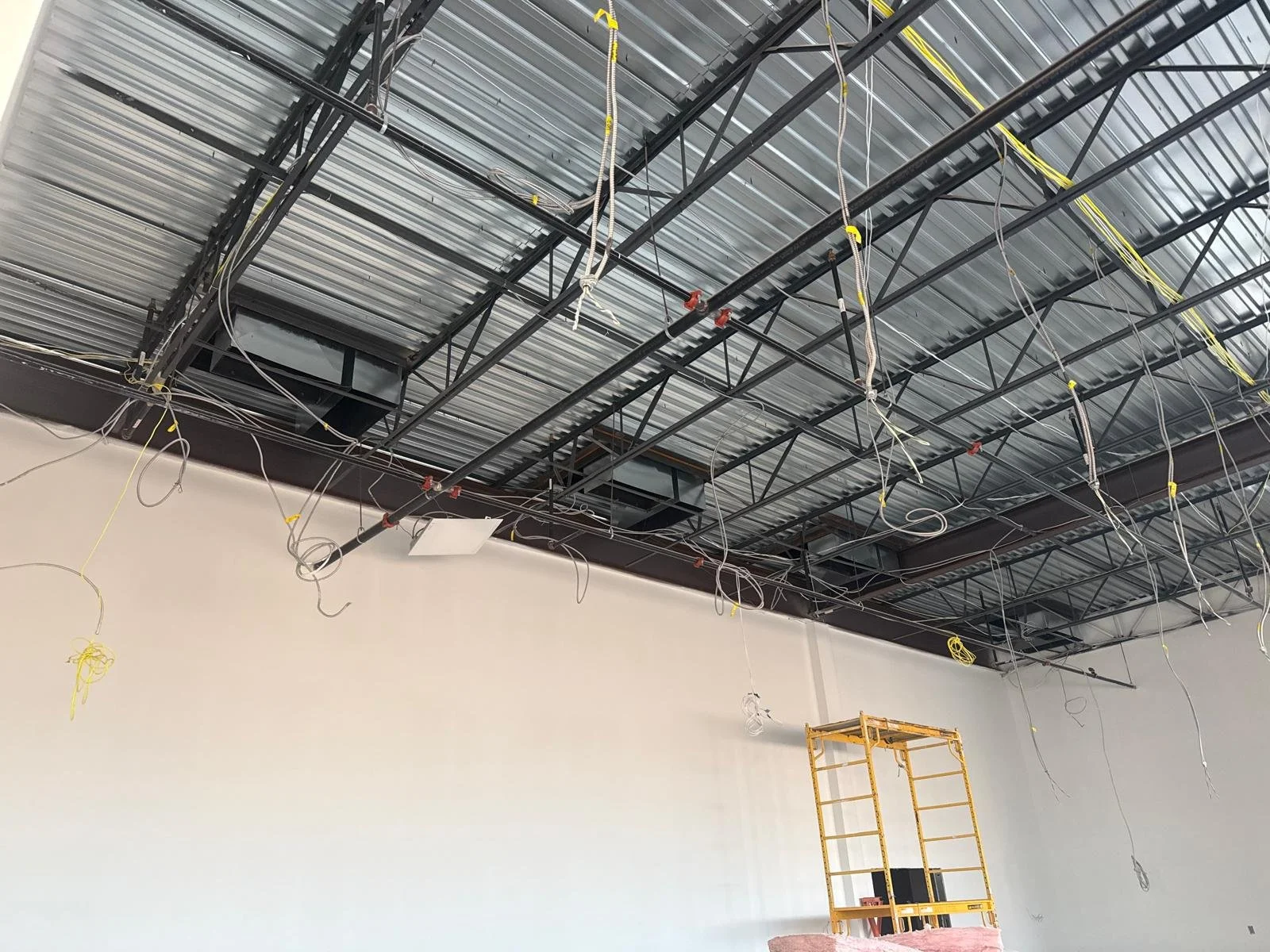 Ceiling of an industrial room under construction with exposed metal framework, wiring hanging down, and a yellow scaffold near the wall.