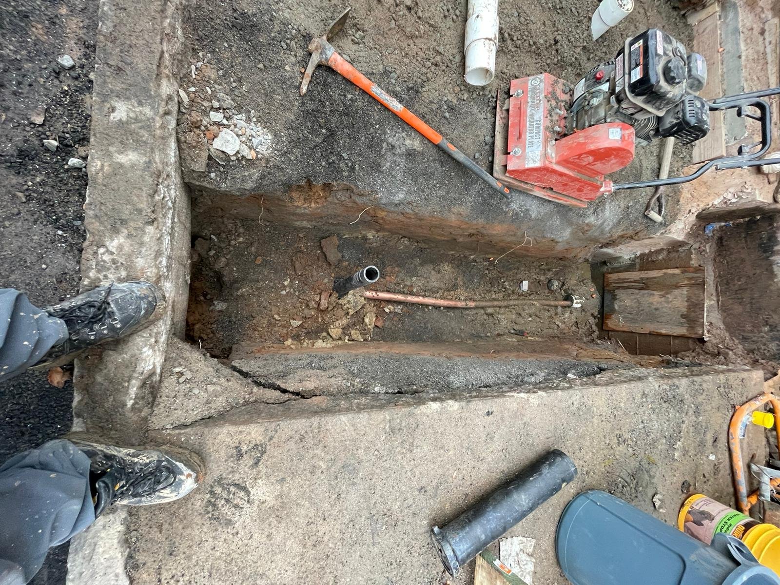 An outdoor construction site showing a trench in the ground with plumbing pipes, construction tools, and equipment around it, including a jackhammer, a pipe, and a worker’s boots.