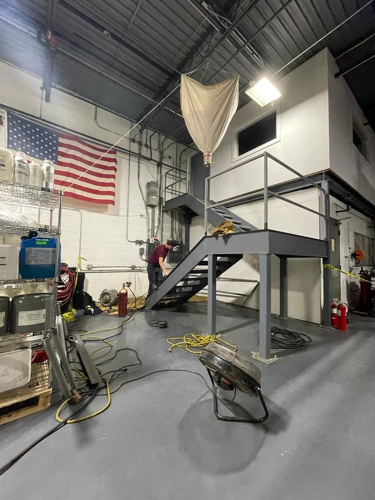 Industrial mezzanine with a staircase, an American flag on the wall, various electrical and safety equipment, and a person working near the wall.
