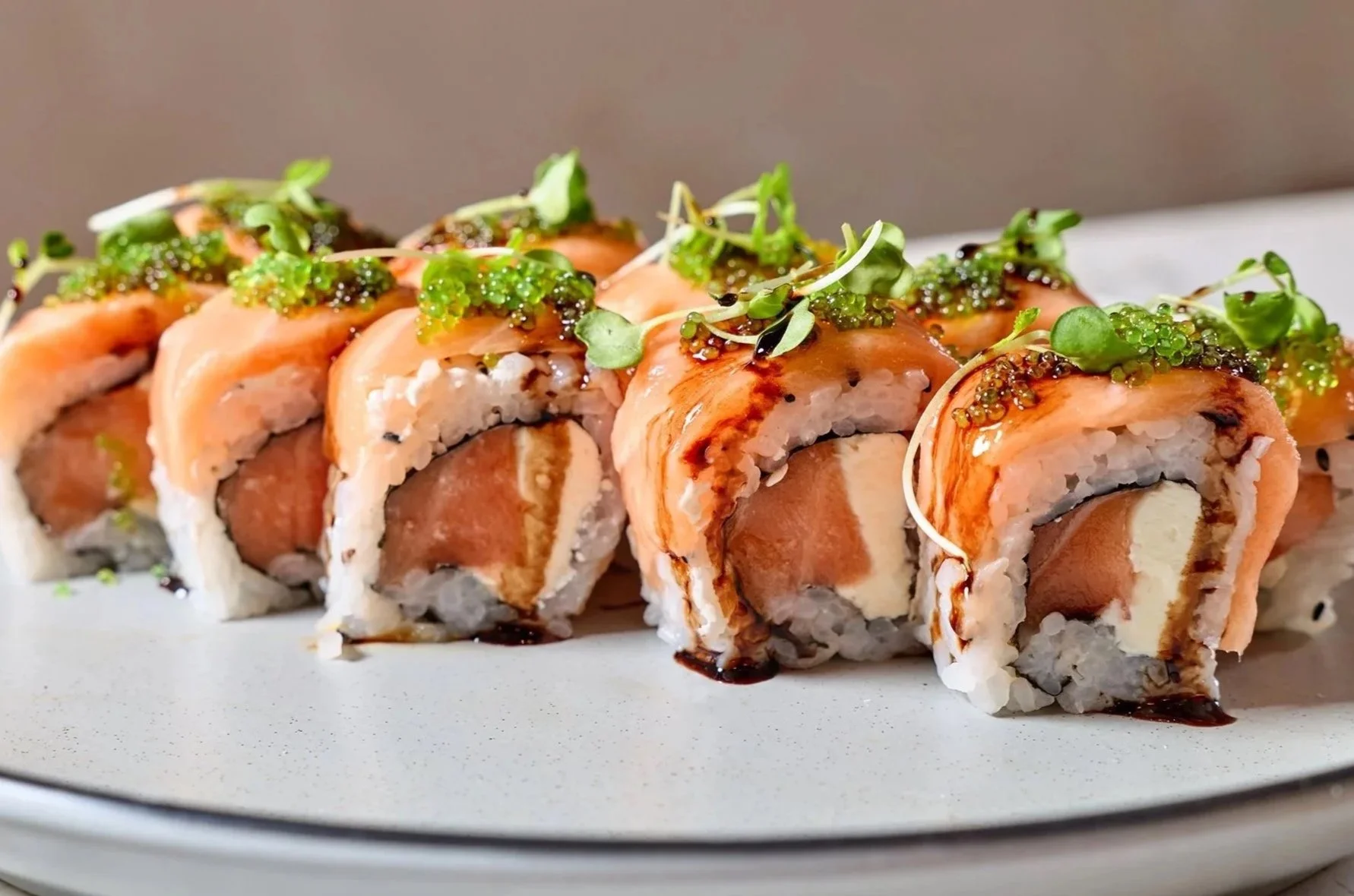 Close-up of sushi rolls topped with microgreens and drizzled with sauce on a white plate.