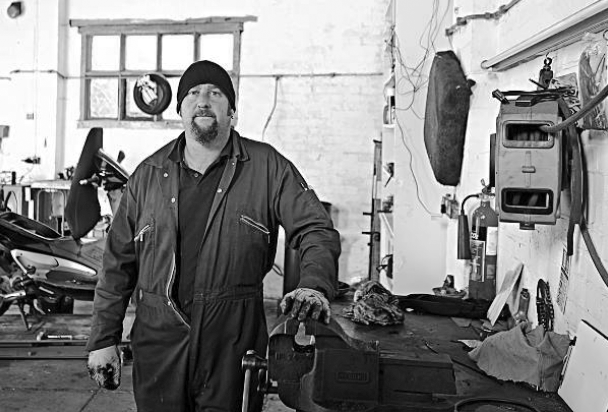 A man in a black beanie and work coveralls standing in a workshop with tools and equipment hanging on the wall behind him, and a workbench with various objects.