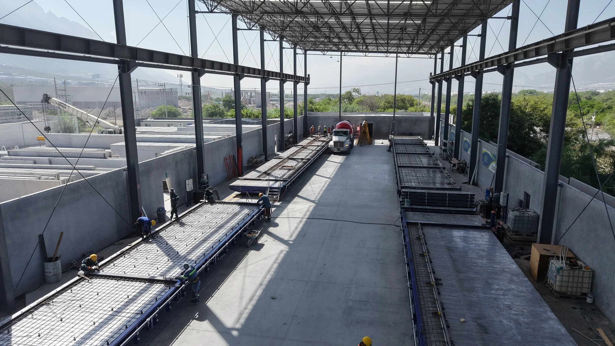 Construction site inside a partially built structure with large open space, workers installing concrete and steel reinforcements, with construction equipment and vehicles present, and outdoor scenery visible through open sides.