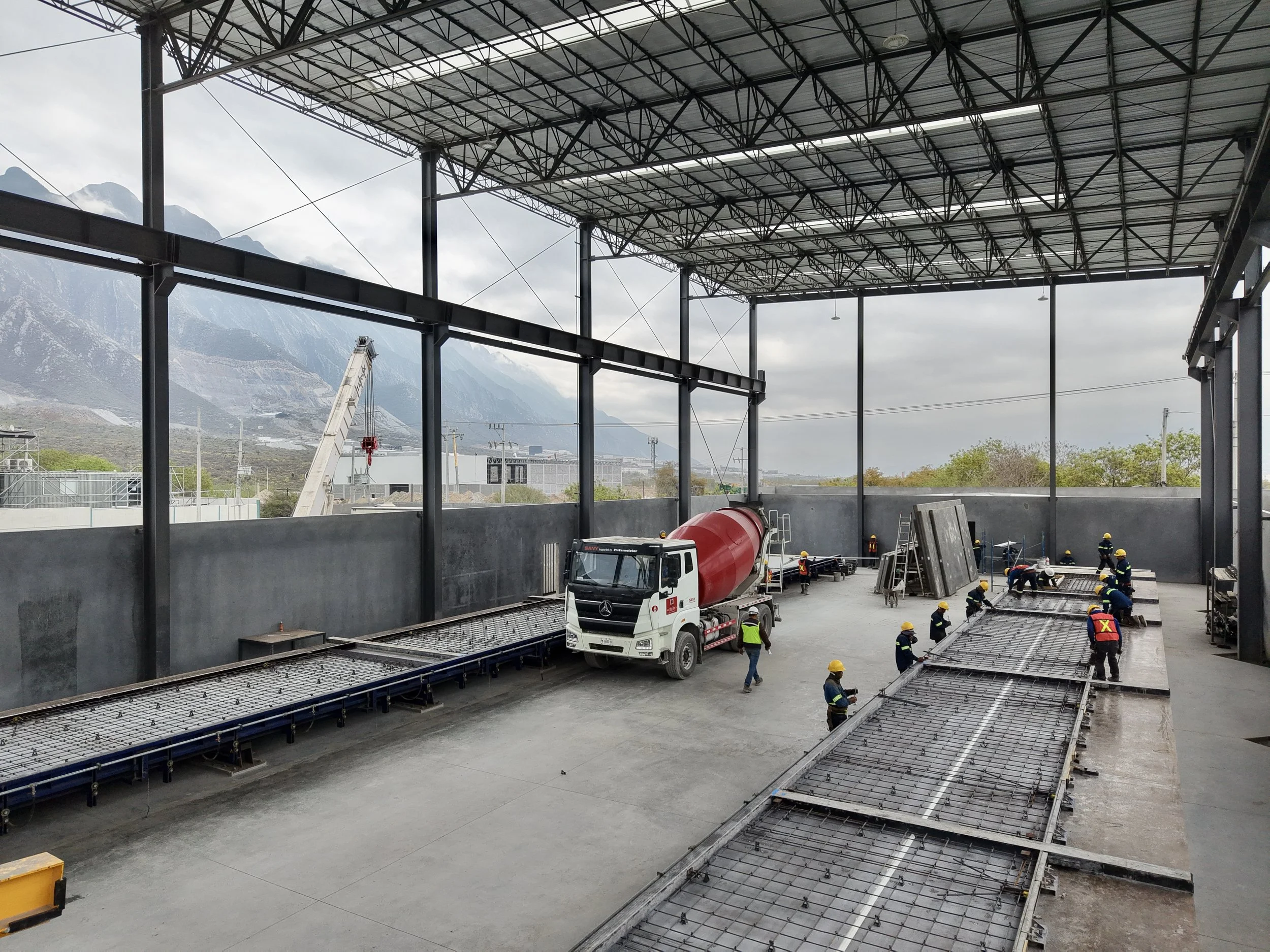 Construction workers installing metal framework inside a large industrial building with an open roof, mountains visible in the background, and a cement mixer truck parked inside.