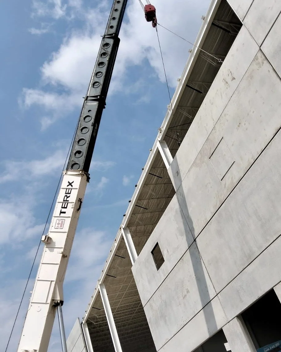 Camión grúa elevando un muro precolado en construcción en un edificio de concreto con cielo azul y nubes.