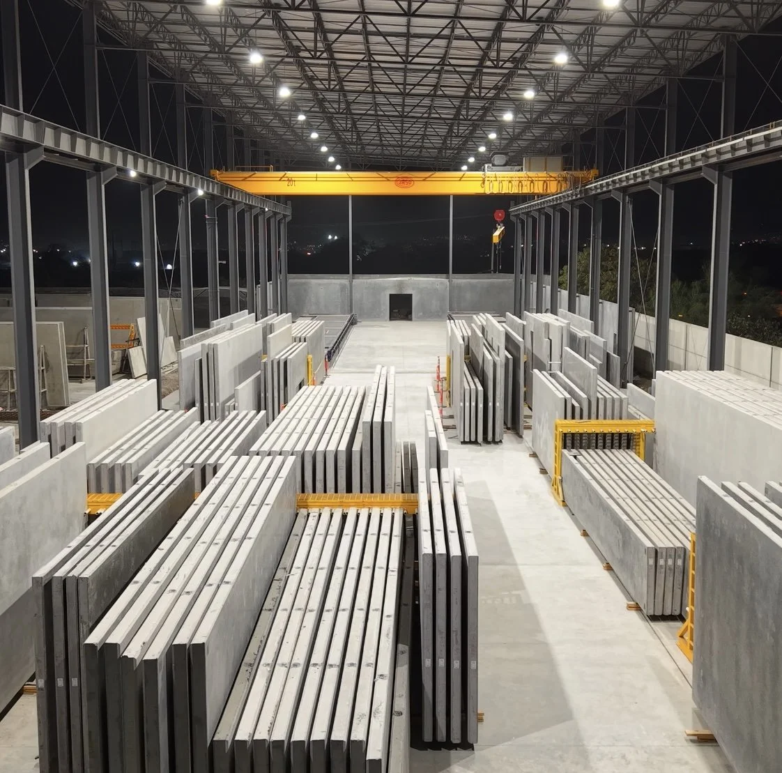 Nighttime view of an industrial warehouse or factory storage area with stacks of metal or concrete slabs organized on the floor, a large yellow overhead crane, and a high ceiling with lighting fixtures.