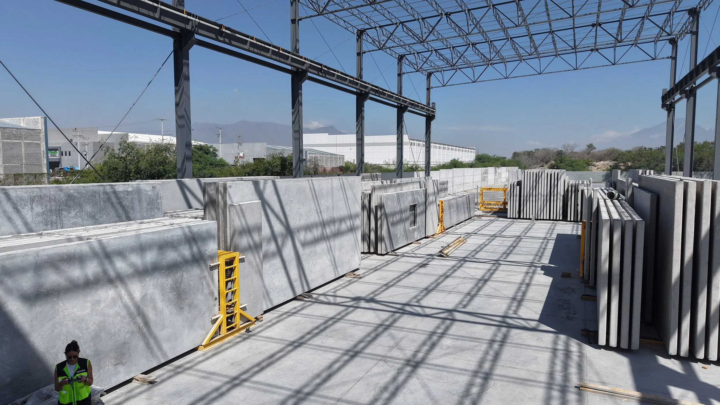 Construction site with large concrete slabs and steel framework under a clear blue sky, with a person in a safety vest in the lower left corner.