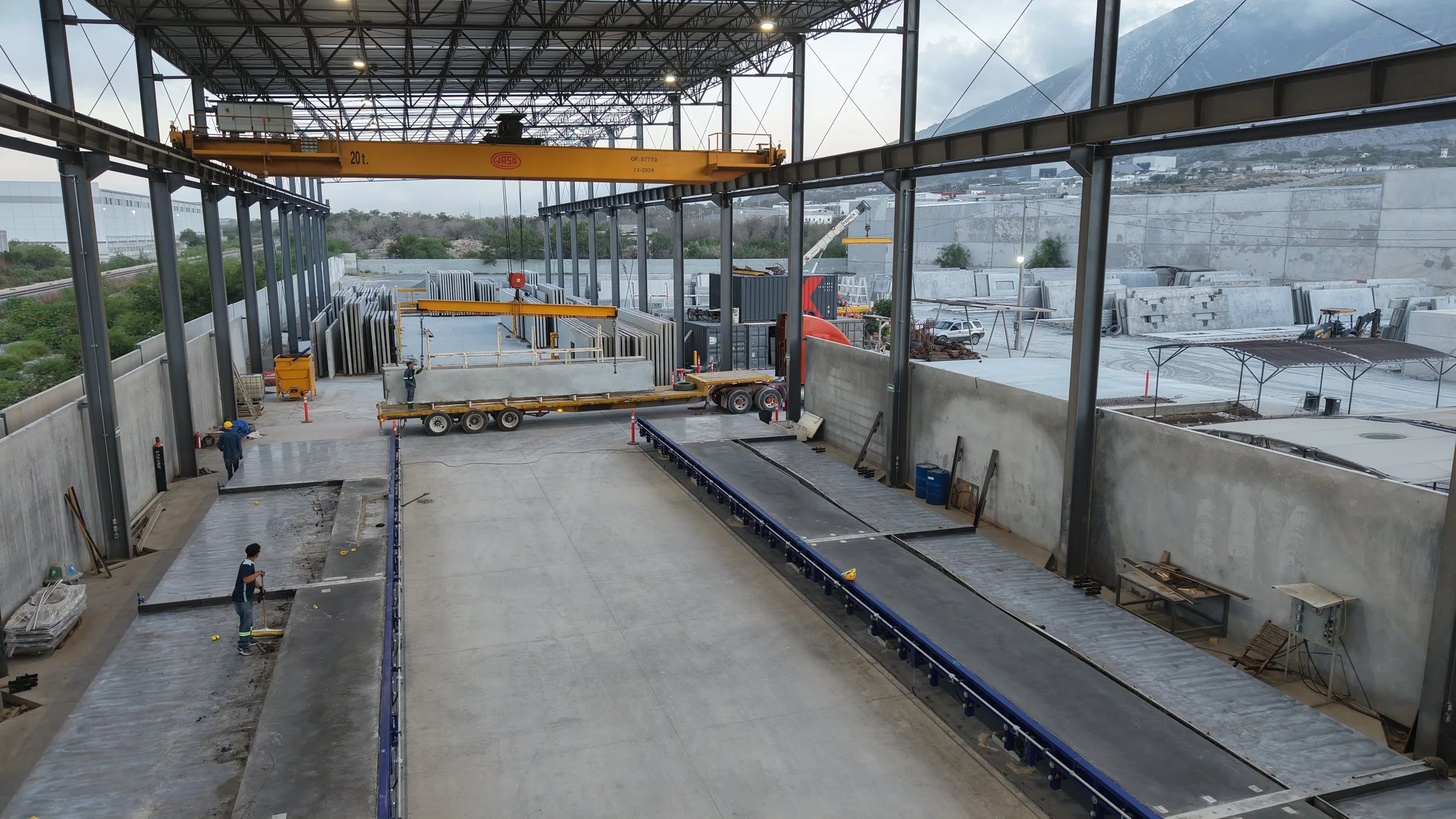 Interior of a large industrial warehouse under construction with workers installing flooring, a yellow overhead crane, and concrete slabs stacked against the walls.