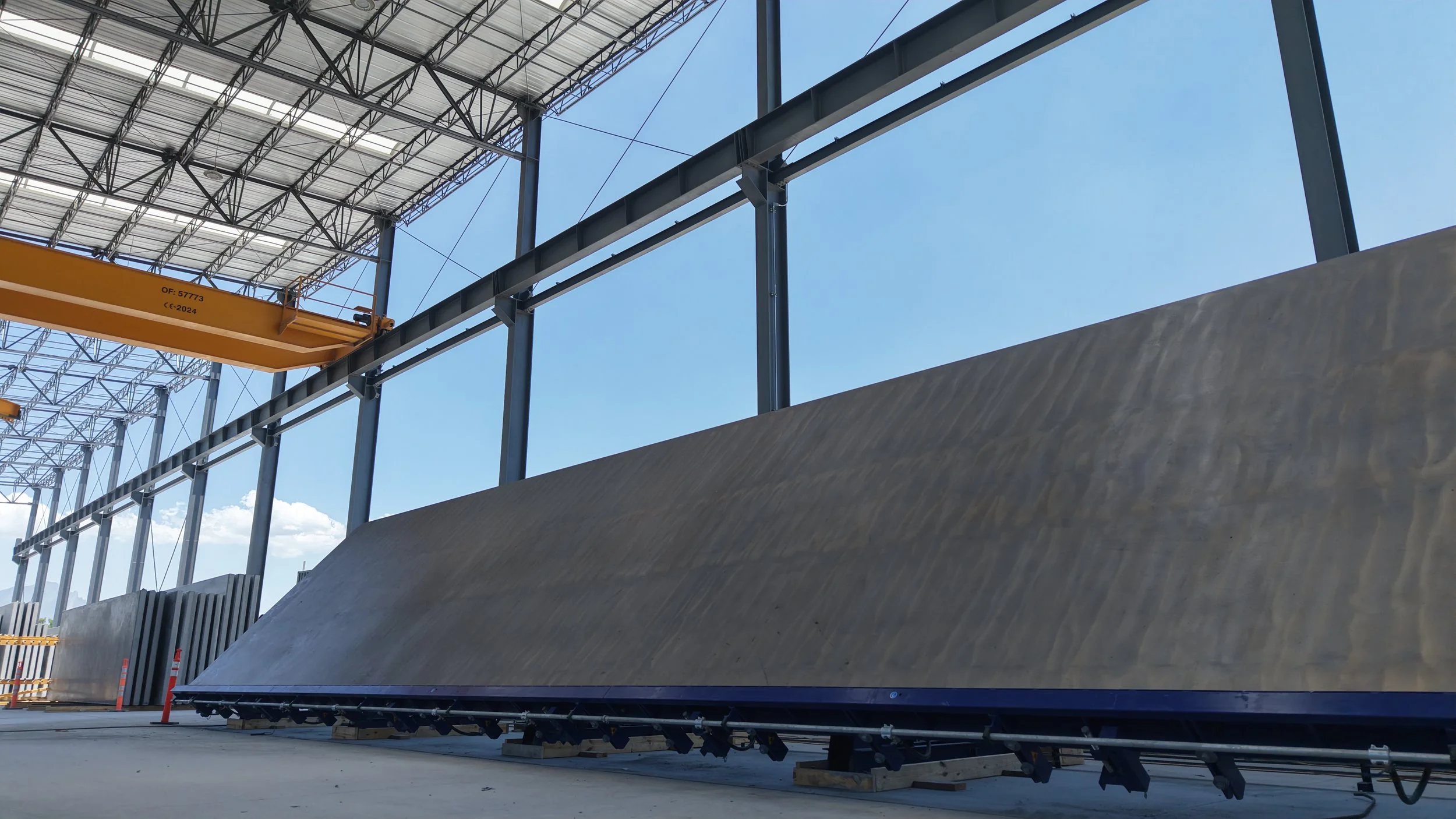 Indoor construction site with a large, angled concrete panel and a yellow overhead crane, under a roof structure with steel framing and skylights.