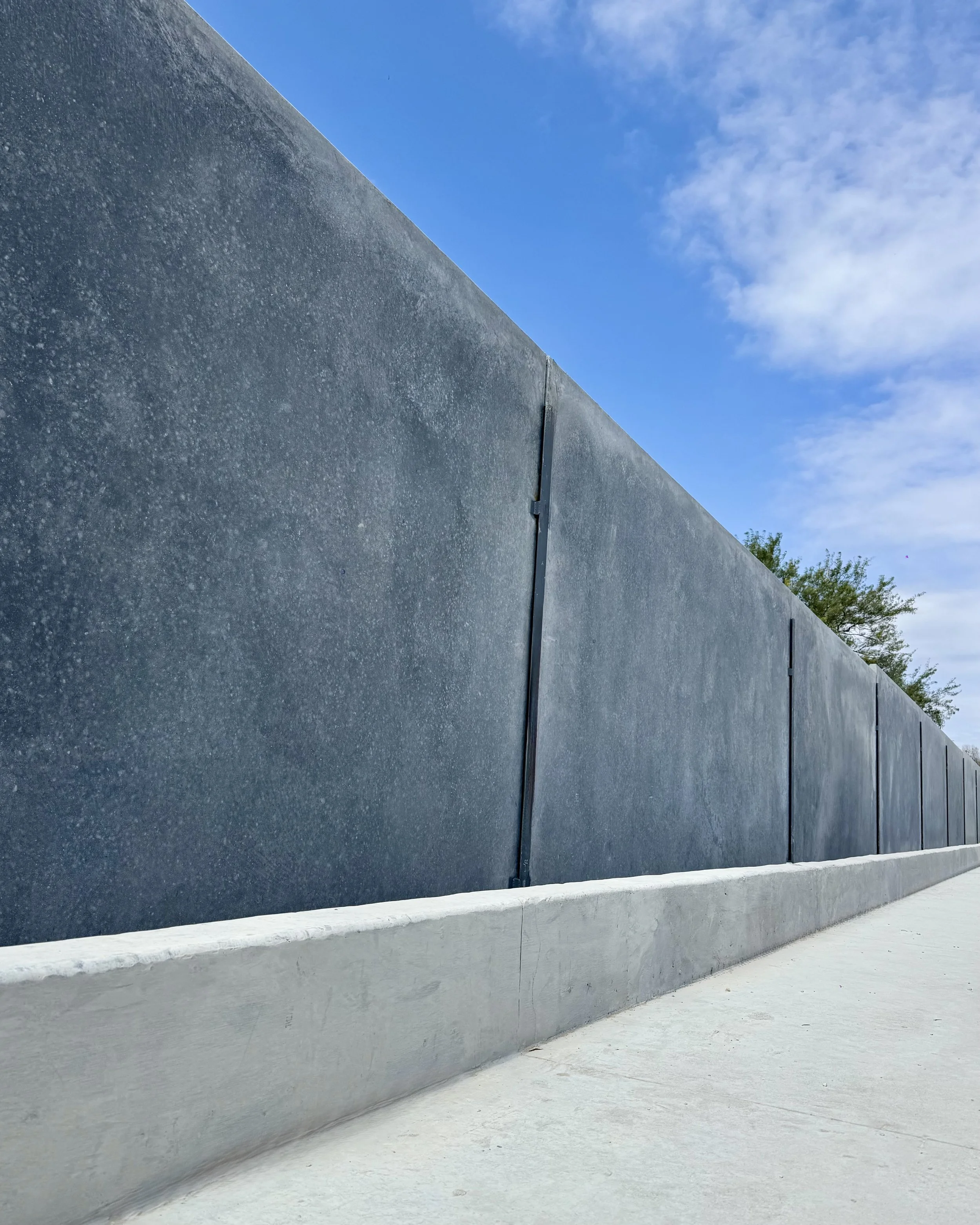 Muro de concreto gris con textura áspera, sobre fondo de cielo azul con nubes dispersas y algunas plantas verdes en la esquina derecha.