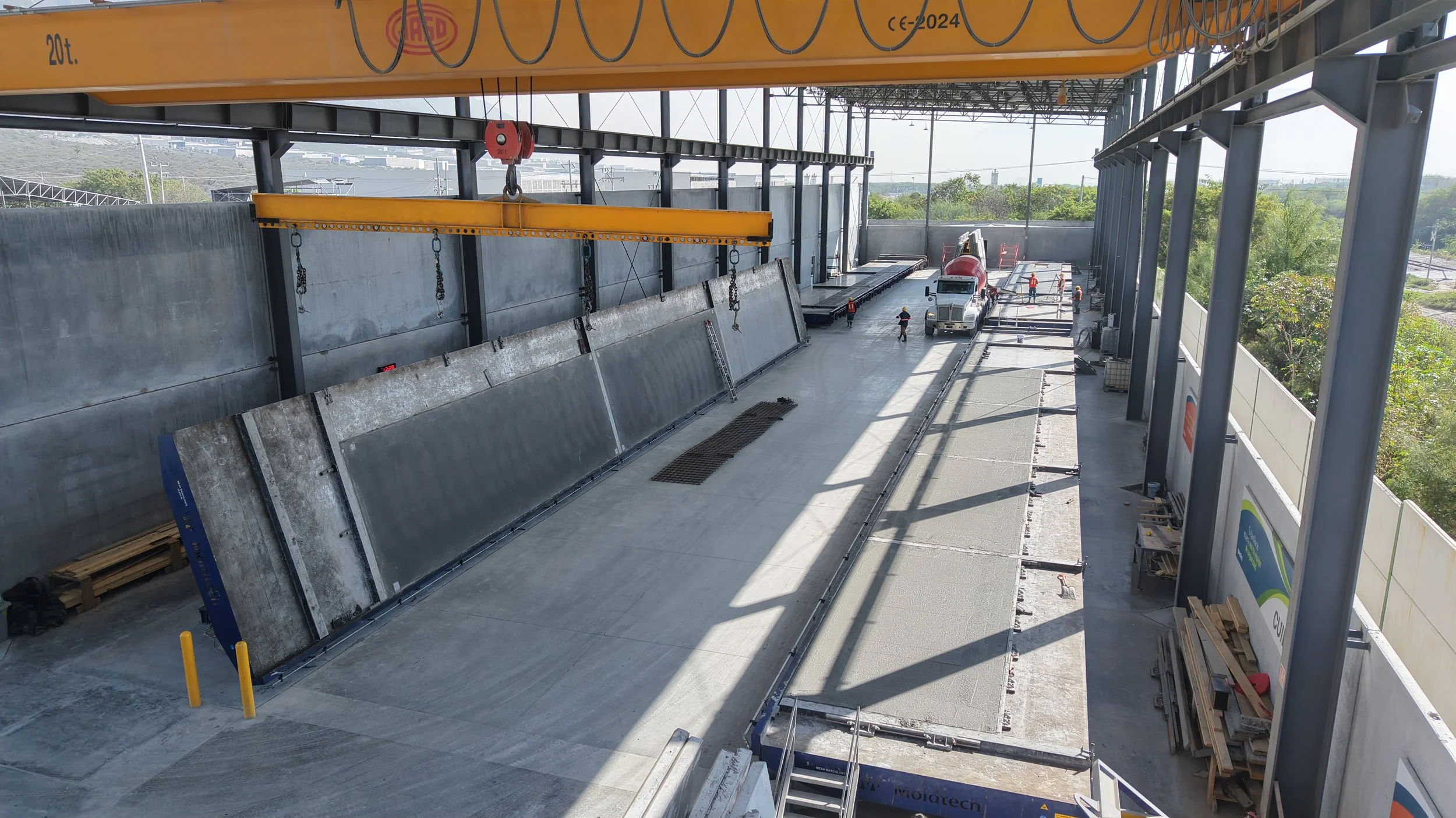 Construction site with large concrete panels being installed on a building's exterior, workers present, a truck and cement mixer in the background, and construction tools and materials on site.