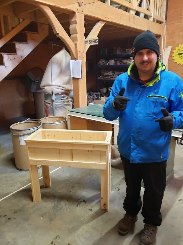 A man in a blue jacket and black beanie giving a thumbs up inside a woodworking or gardening workshop. There is a wooden planter box on the floor in front of him, with tools and supplies like large buckets and a trash can visible in the background.