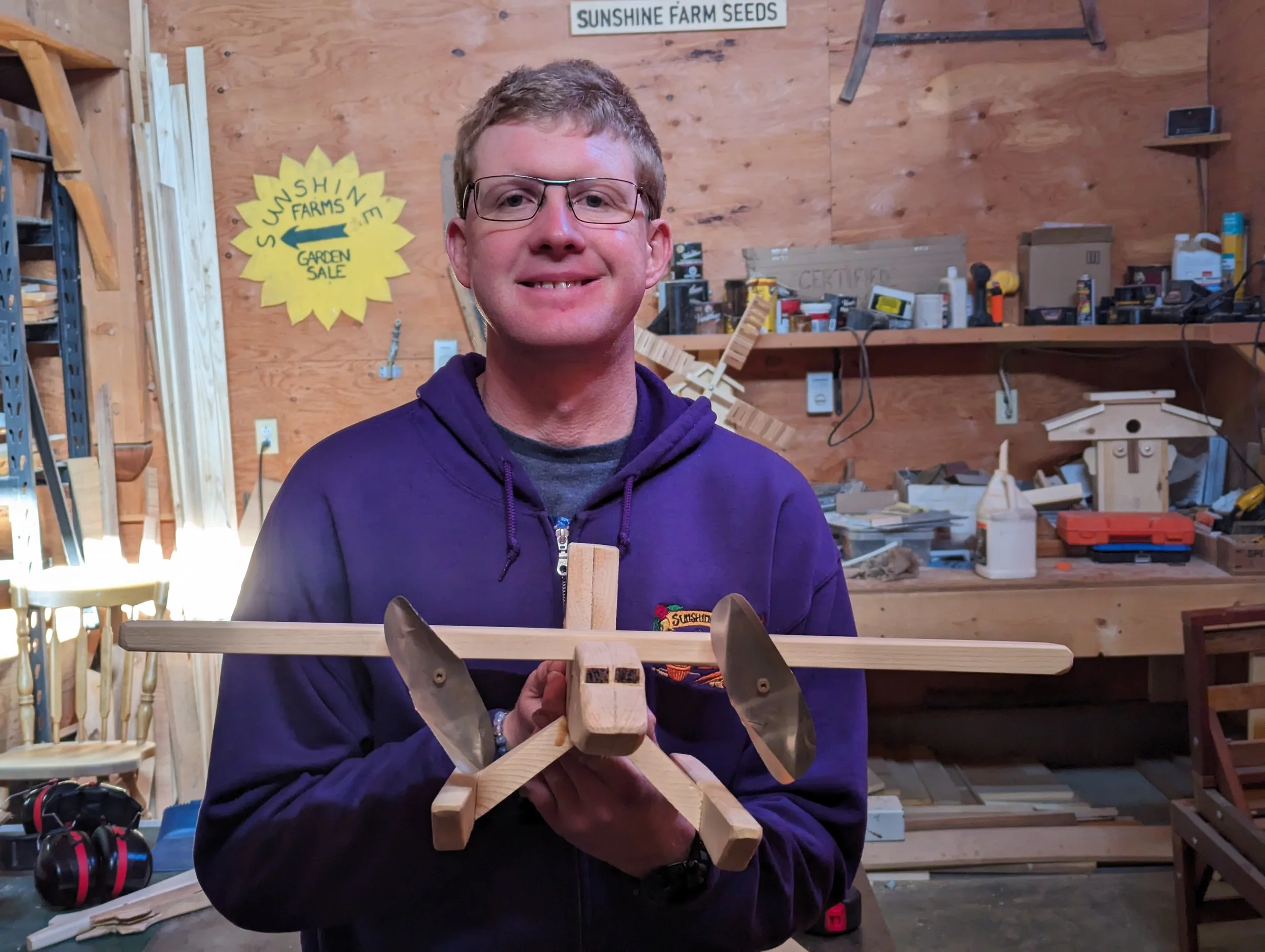 A smiling man with glasses holding a wooden airplane model in a woodworking workshop.