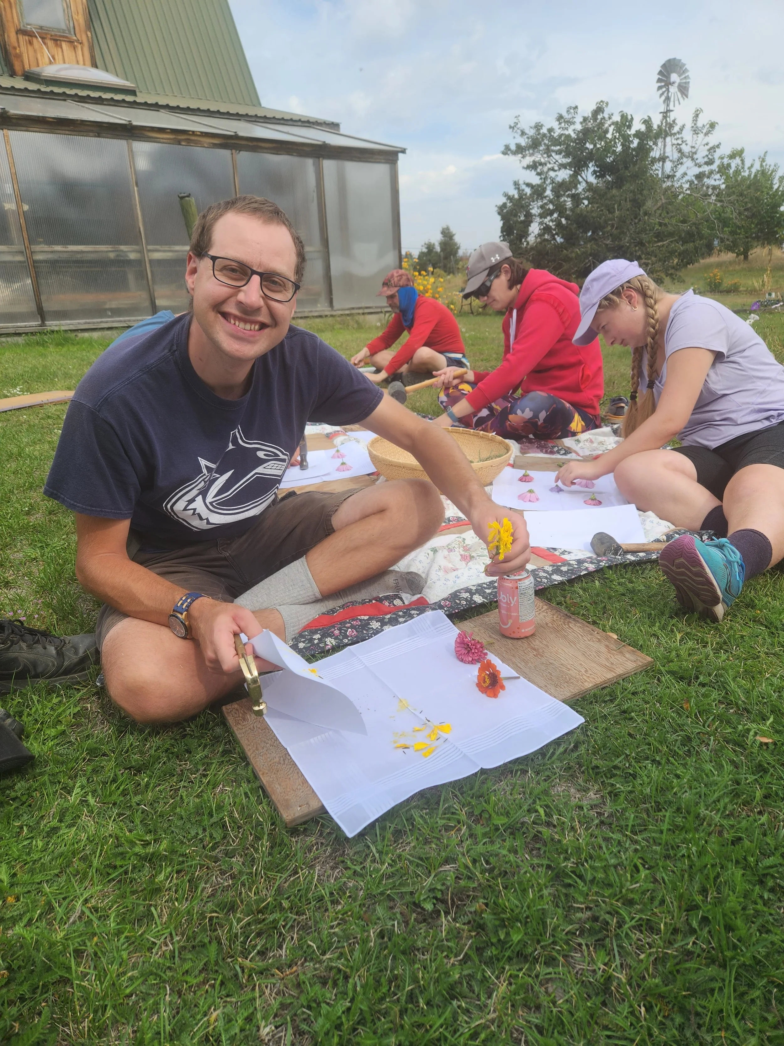 A man sitting on grass, smiling, while working on a craft project with three children in an outdoor garden or farm setting.