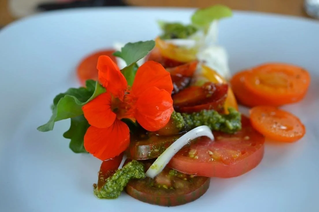 A plated salad with sliced heirloom tomatoes, fresh herbs, edible flowers, and green vegetables, garnished with edible flowers and a small cup of dressing.