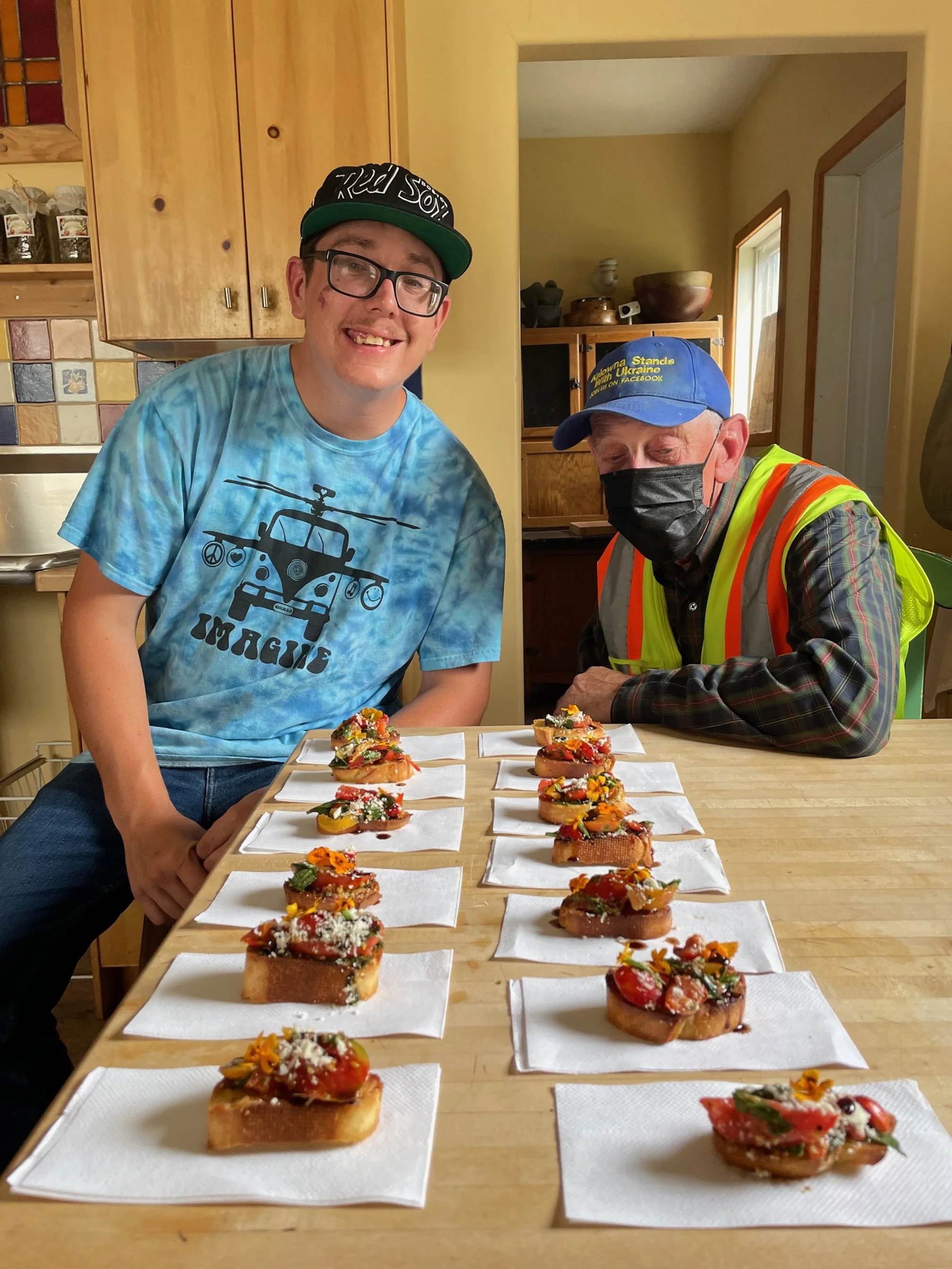 Two men sitting at a kitchen table with colorful topped bread slices on white napkins.