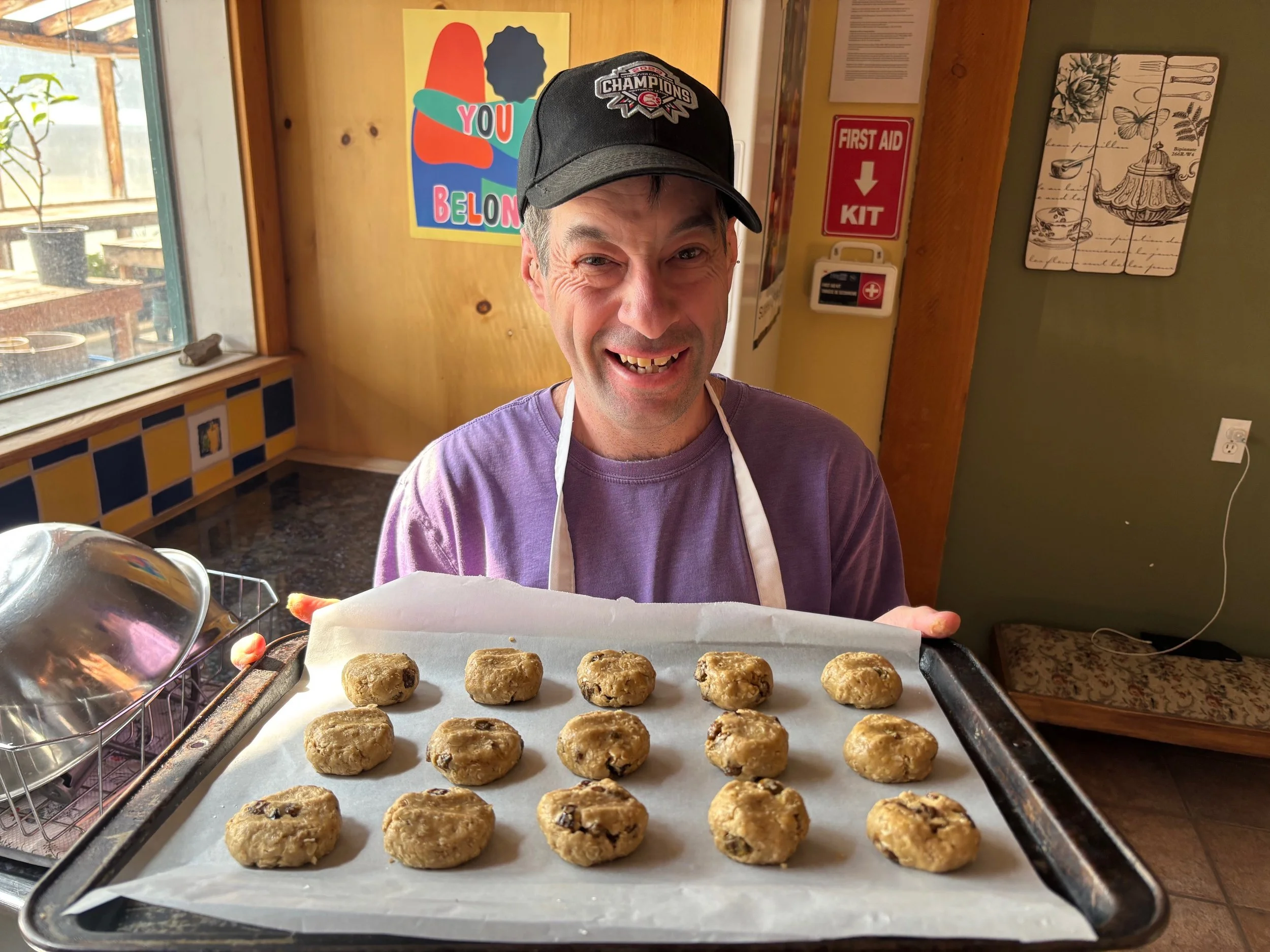 A man wearing a black baseball cap and purple shirt is smiling and holding a tray of freshly baked cookies.