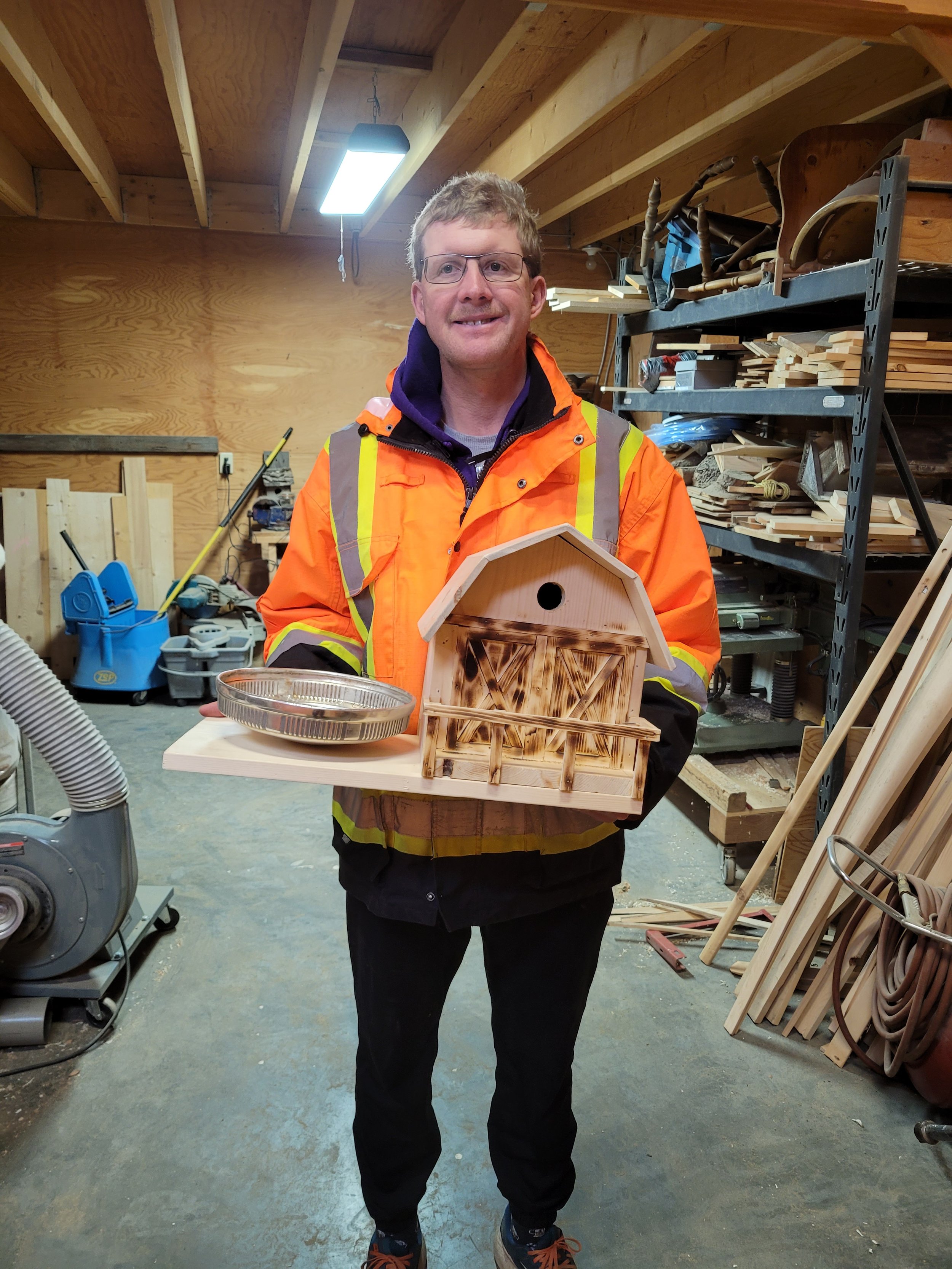 A man in a brightly colored safety jacket holding a small wooden birdhouse and a metal tray inside a woodworking shop.