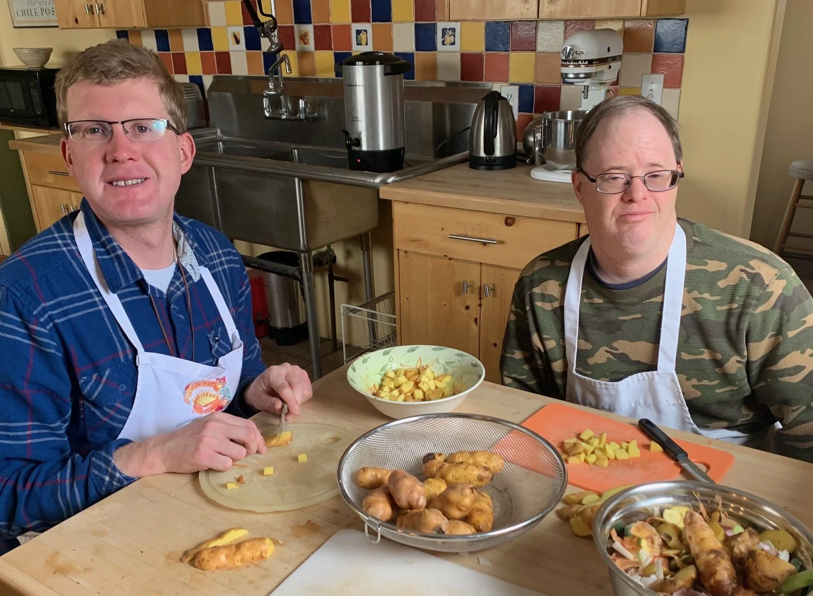 Two men in aprons sit at a kitchen table preparing food. One man is chopping a yellow vegetable, possibly ginger, while the other is sitting next to a cutting board with chopped food. There are bowls of peeled baby potatoes and other ingredients on t