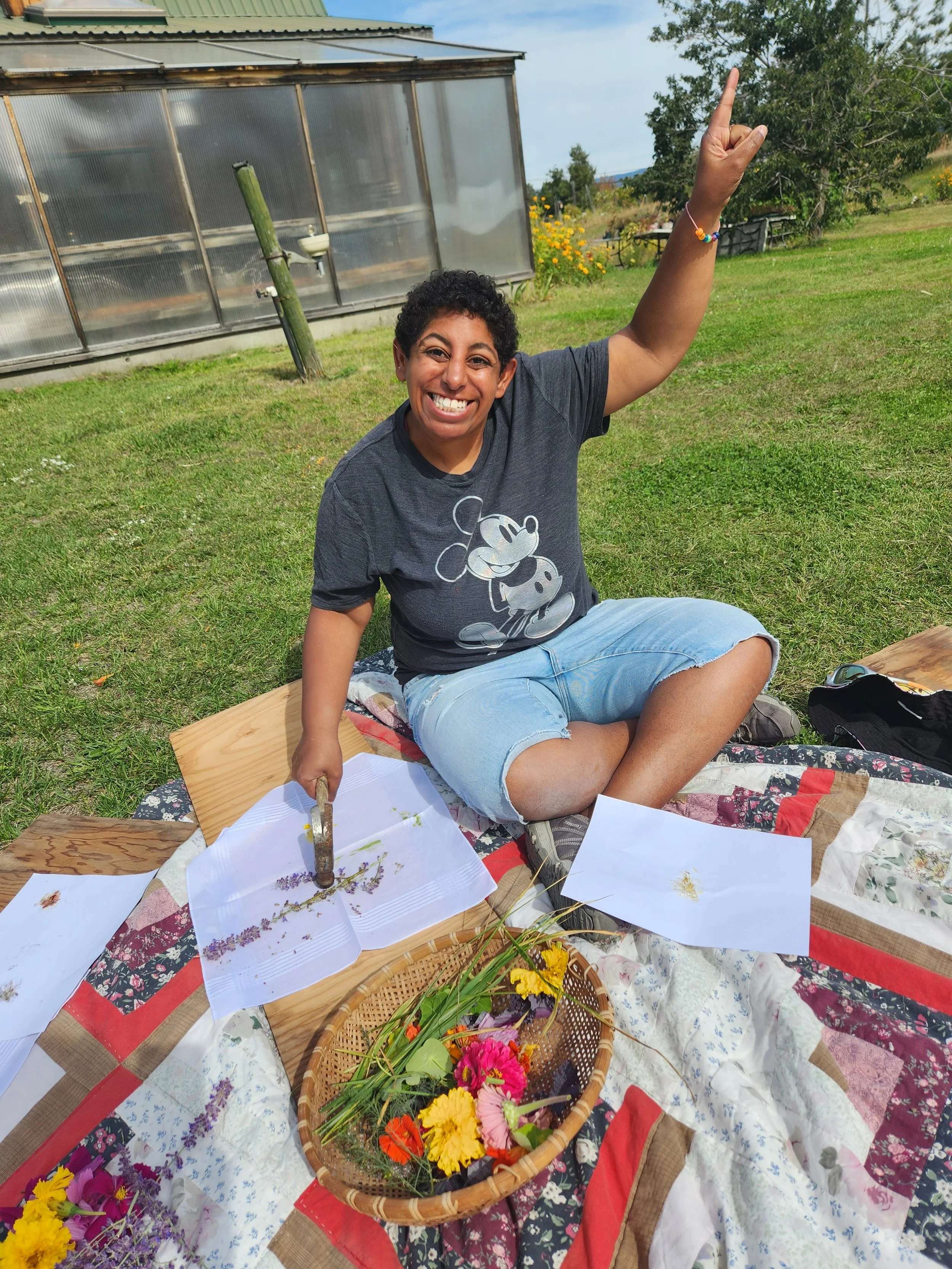 A woman sitting on a quilt outdoors, raising one arm with her index finger up, smiling brightly, surrounded by flowers and papers, with greenery and a greenhouse in the background.