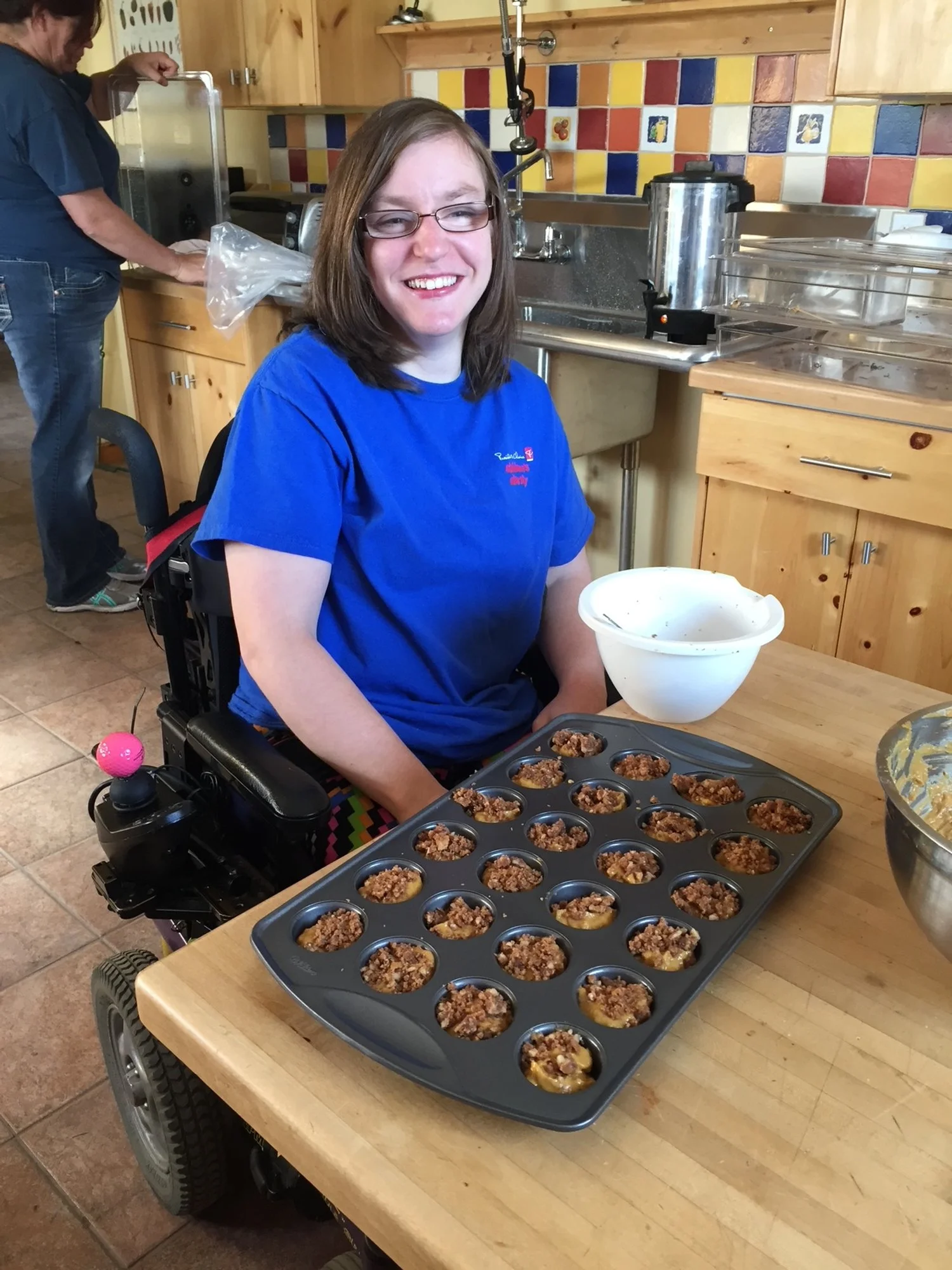A smiling woman in a blue shirt sitting at a kitchen table with a tray of mini pies topped with crumbly streusel. There is a mixing bowl and a bowl of filling on the table. In the background, a person is working in the kitchen.