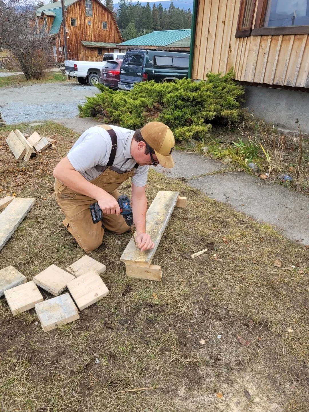 A man kneeling on grass and working with a power drill on a piece of wood outdoors near a house, with cars and shrubs in the background.