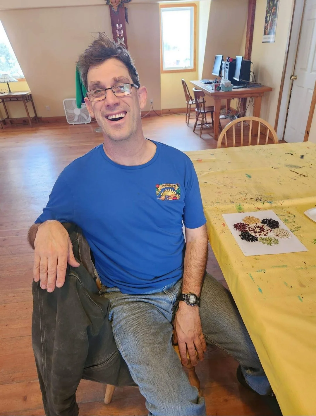 A man with glasses, smiling, sitting at a table with a yellow tablecloth, with various beans on a piece of paper nearby. He is wearing a blue t-shirt and gray pants, inside a room with wooden floors and furniture.