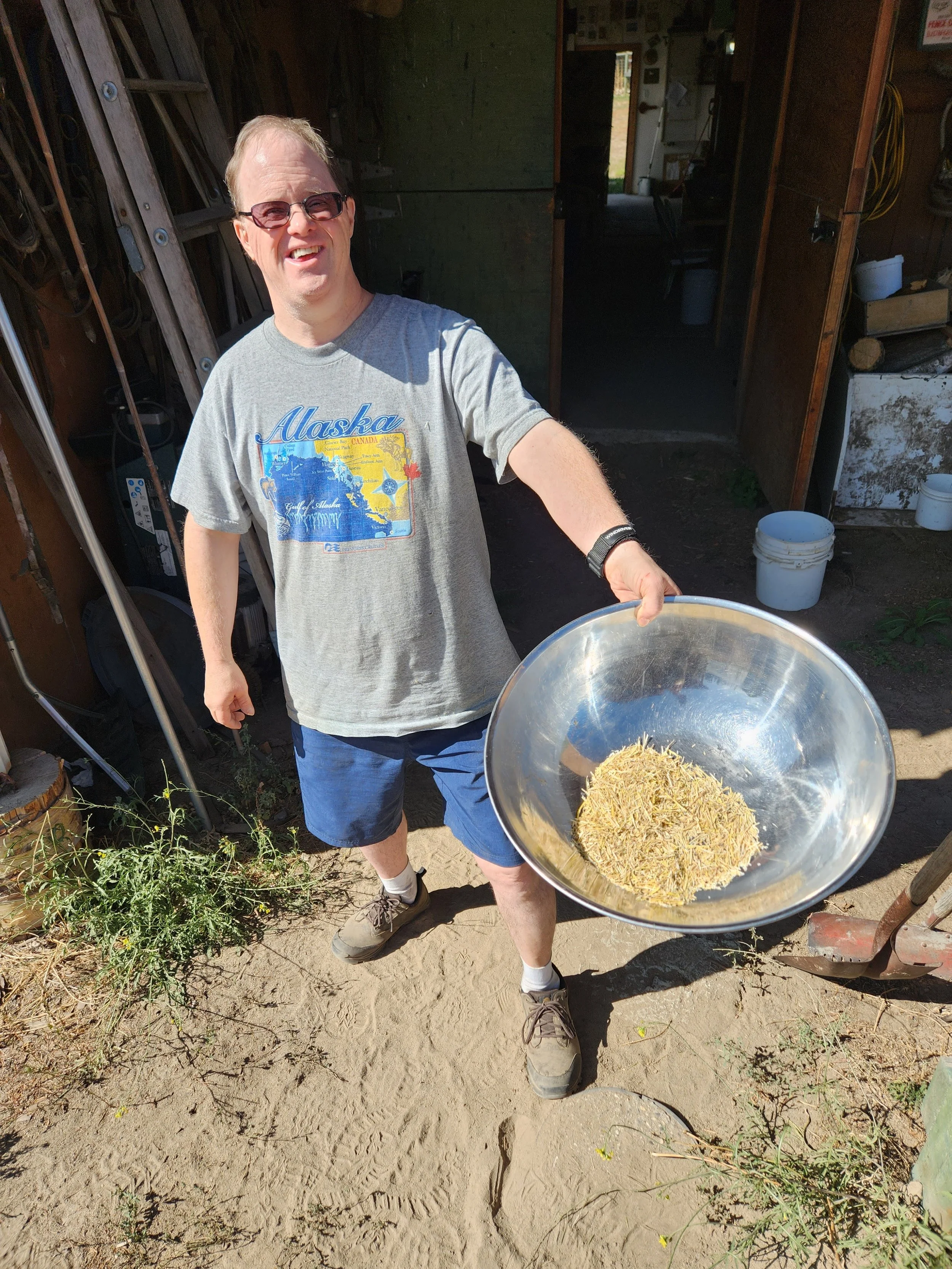 A man holding a metal bowl filled with dried herbs, standing outdoors near a shed with gardening tools and buckets visible in the background.