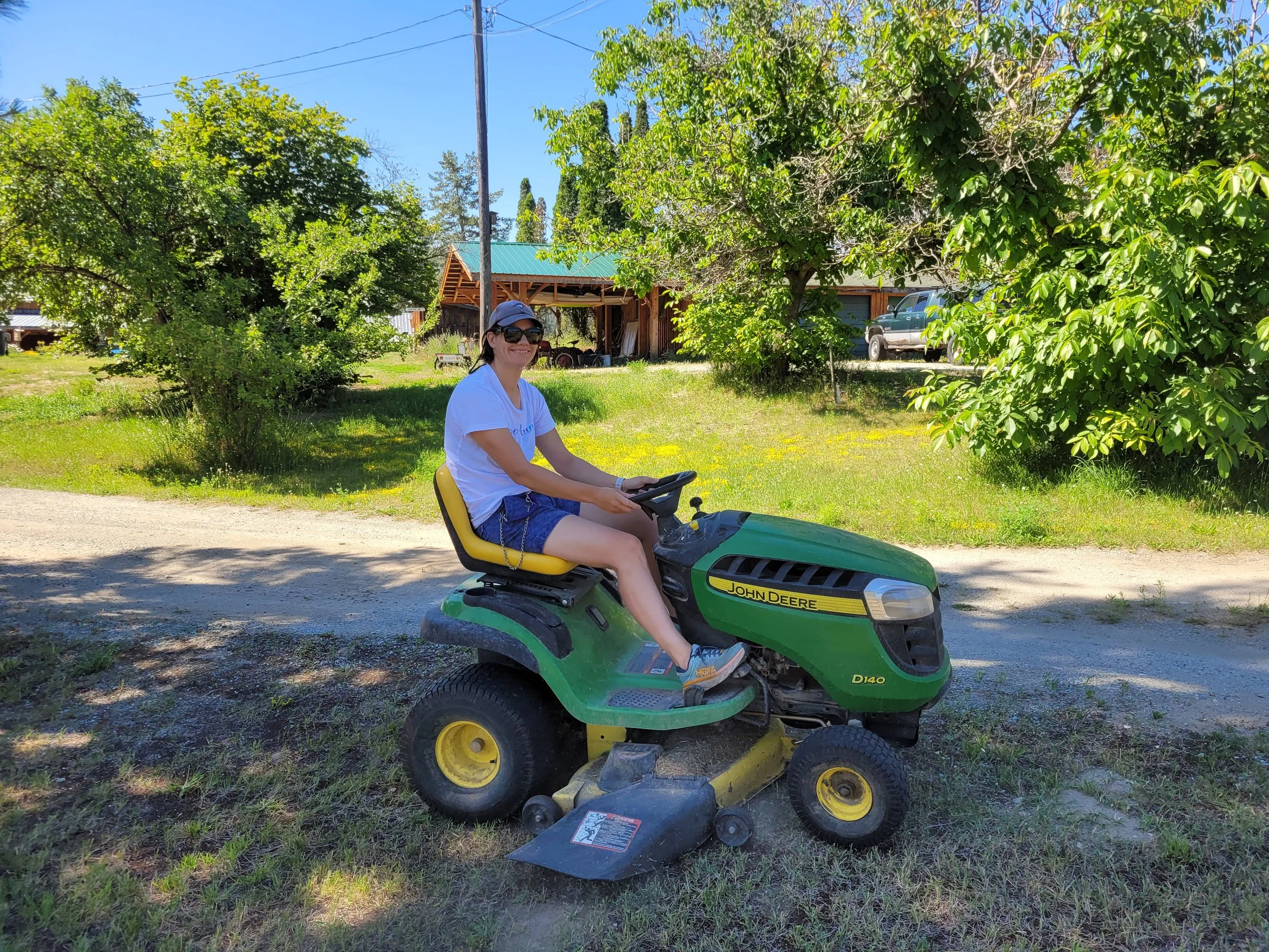 A woman sitting on a green and yellow John Deere riding lawn mower outdoors on a sunny day.