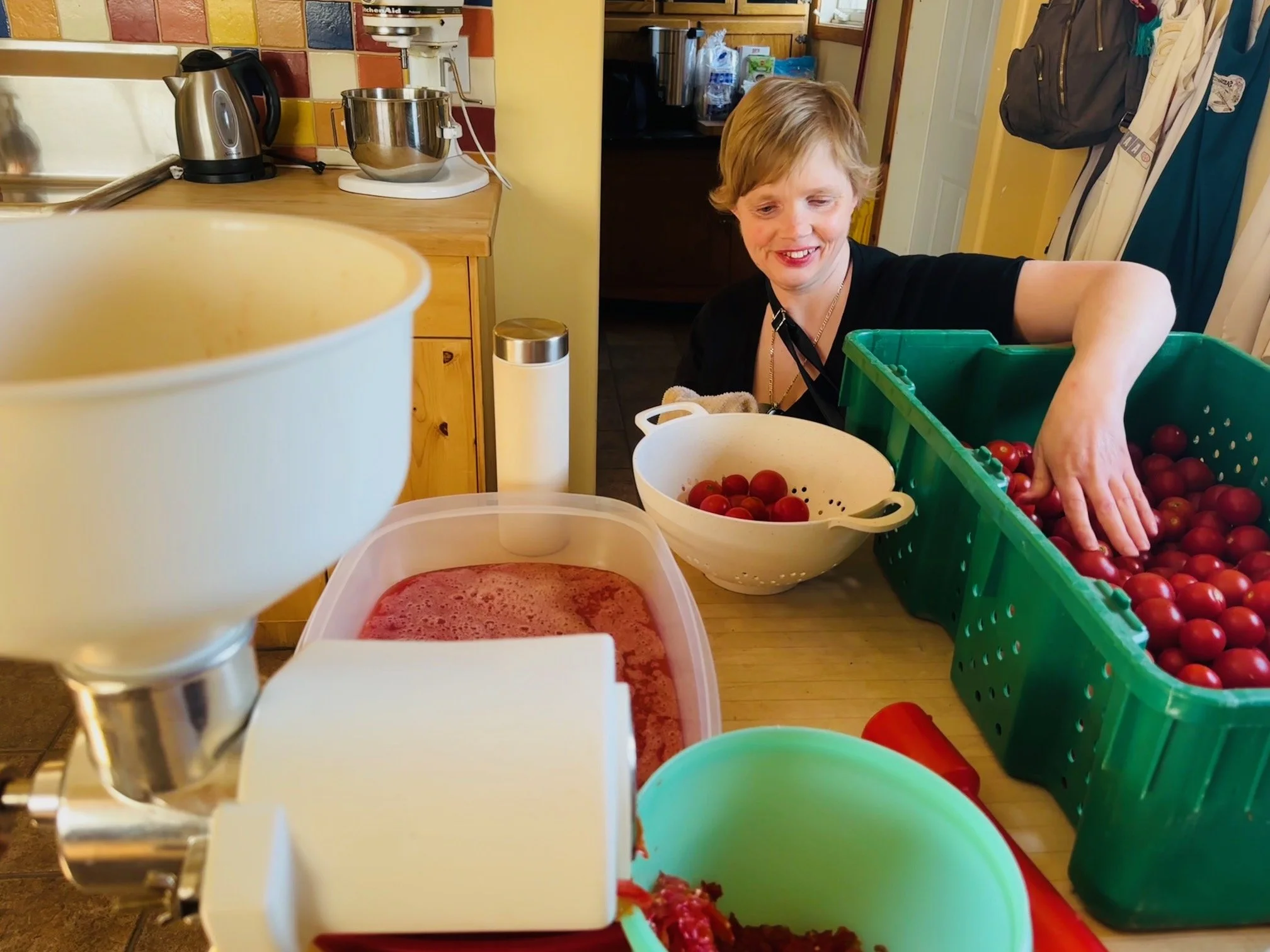 A woman smiling and reaching into a green basket full of red cherry tomatoes in a kitchen, with various kitchen appliances and containers on the counter.