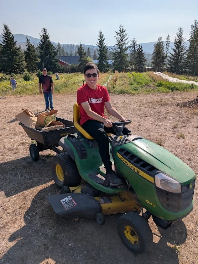A young boy sitting on a green John Deere riding lawn mower with a yellow seat in a rural field, smiling at the camera, with people and trees in the background.