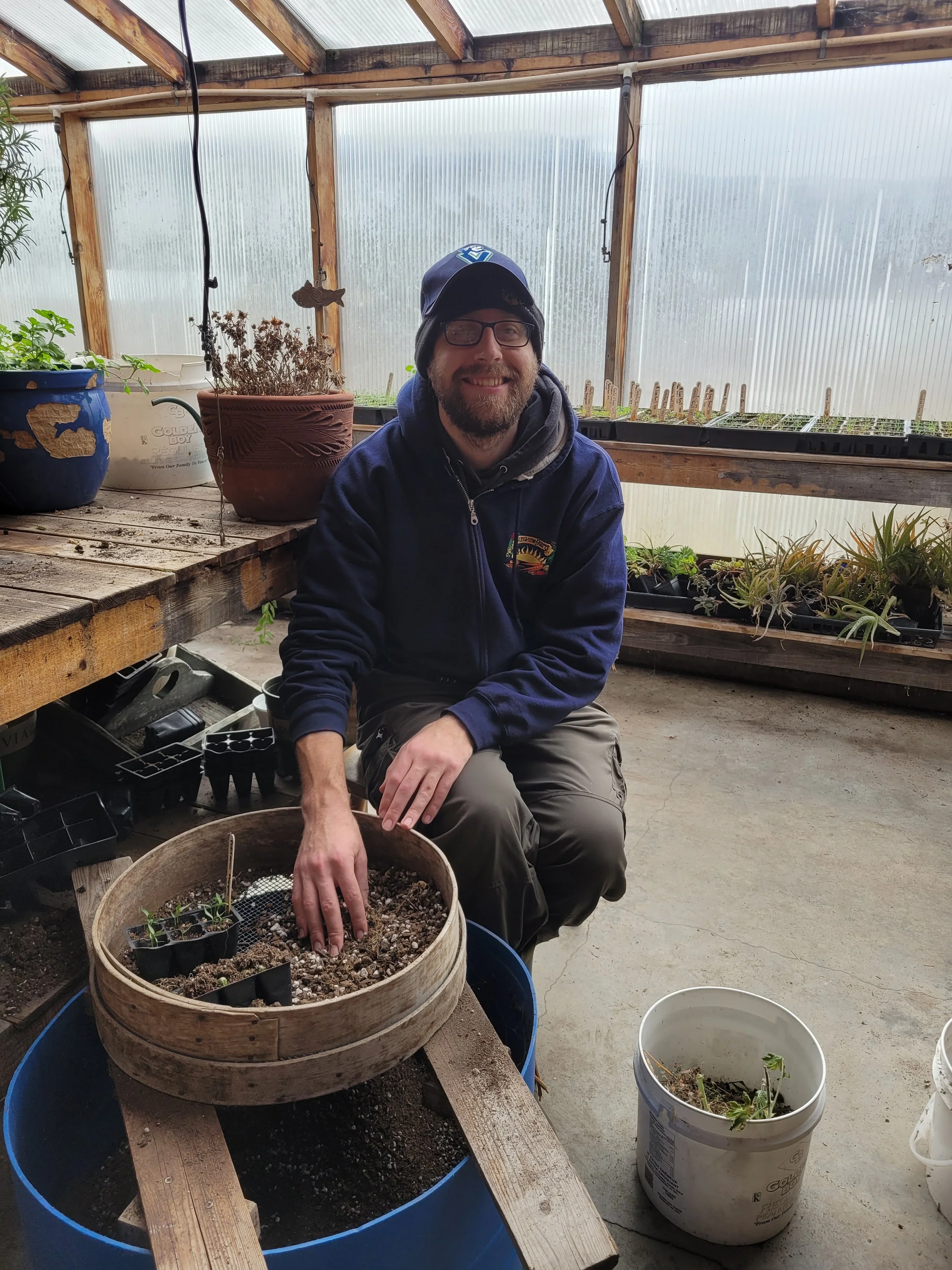 A man smiling while gardening inside a greenhouse, sitting beside a large round wooden container filled with soil and small plants, surrounded by flower pots and gardening supplies.