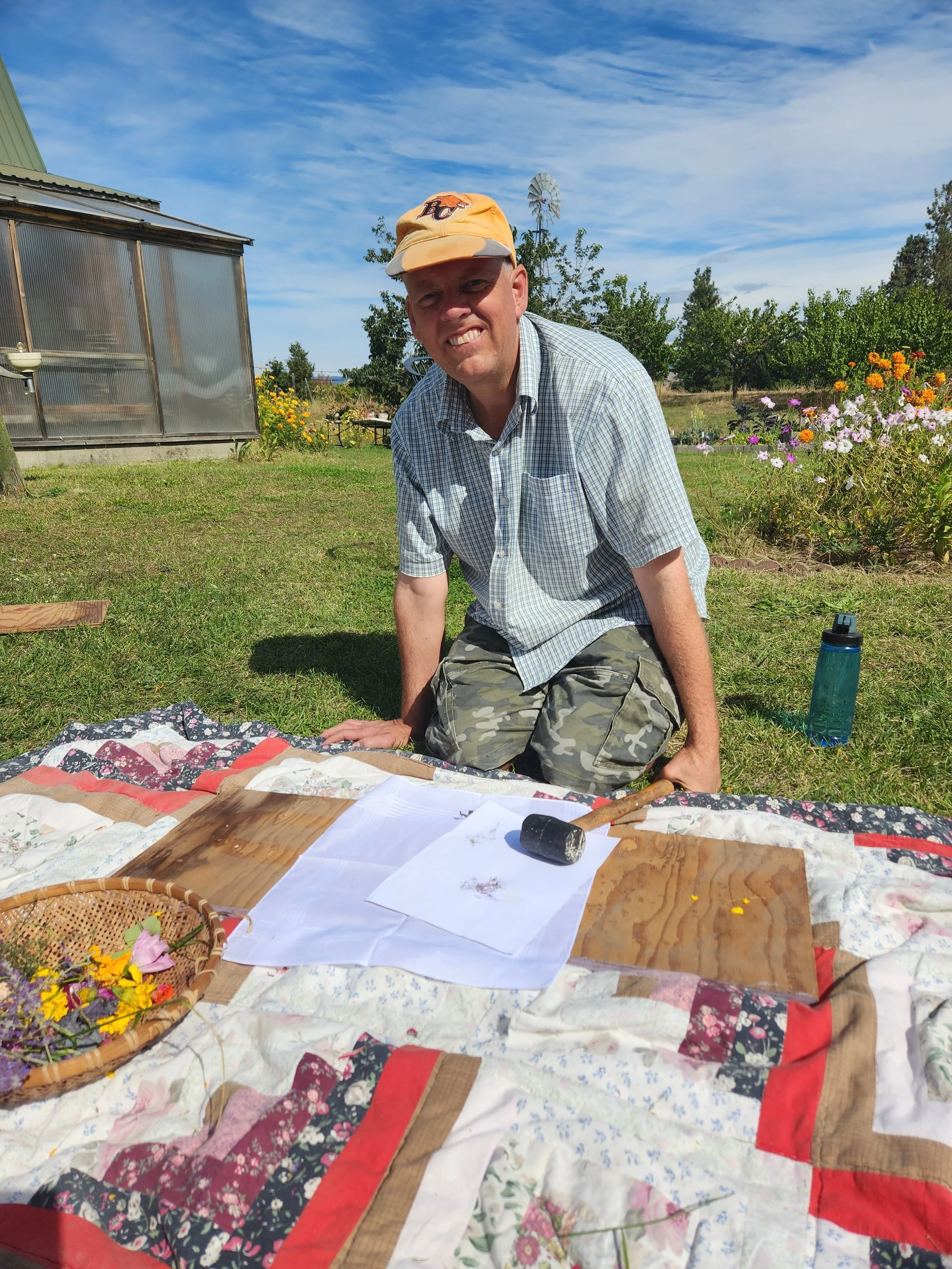 Man kneeling on grass outdoors in a sunny garden, surrounded by flowers and a greenhouse, with a wooden mallet, papers, and a basket of flowers on a quilted blanket.