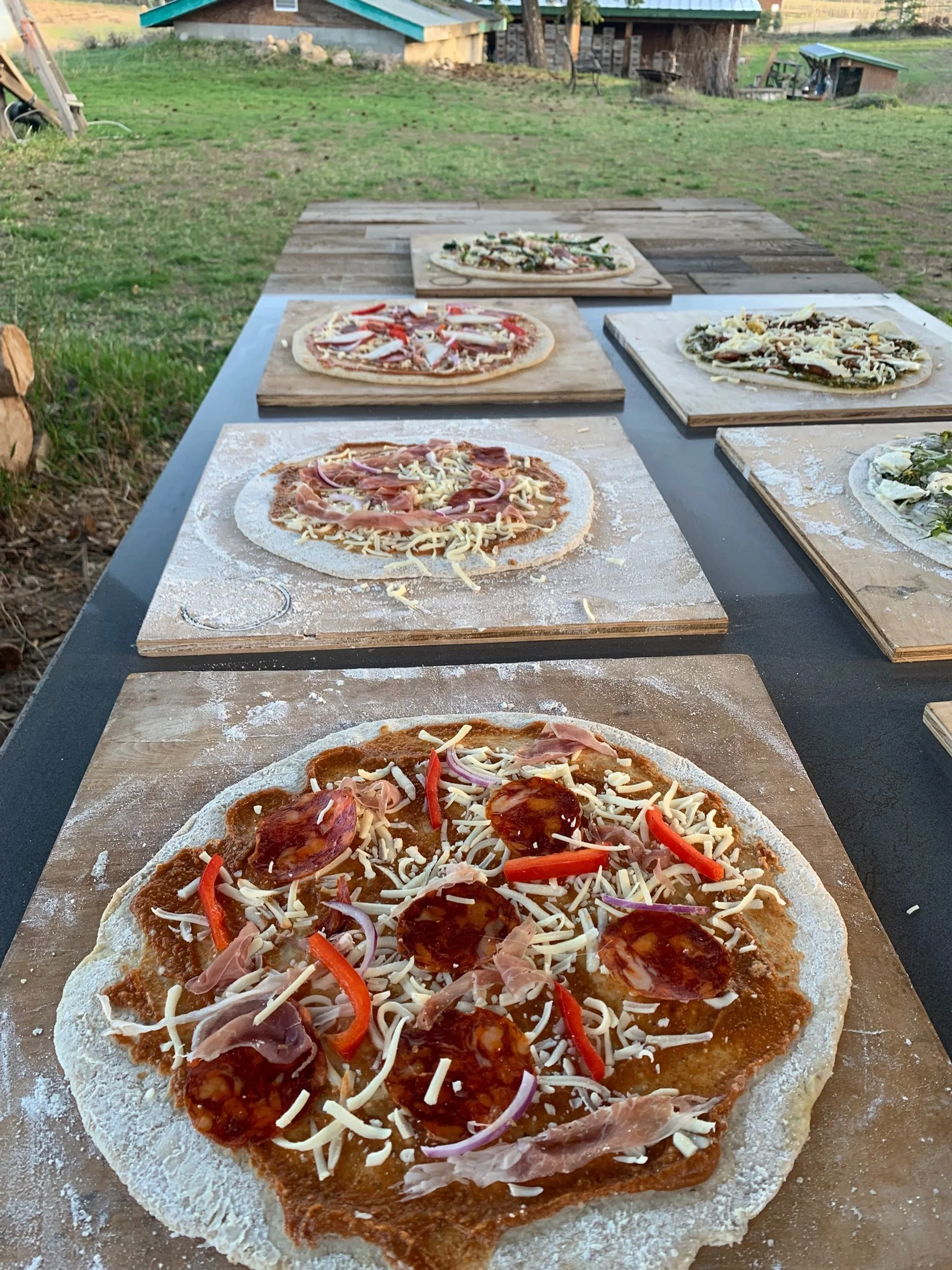 Several uncooked pizzas with toppings like cheese, pepperoni, onions, and red peppers on wooden boards outdoors.