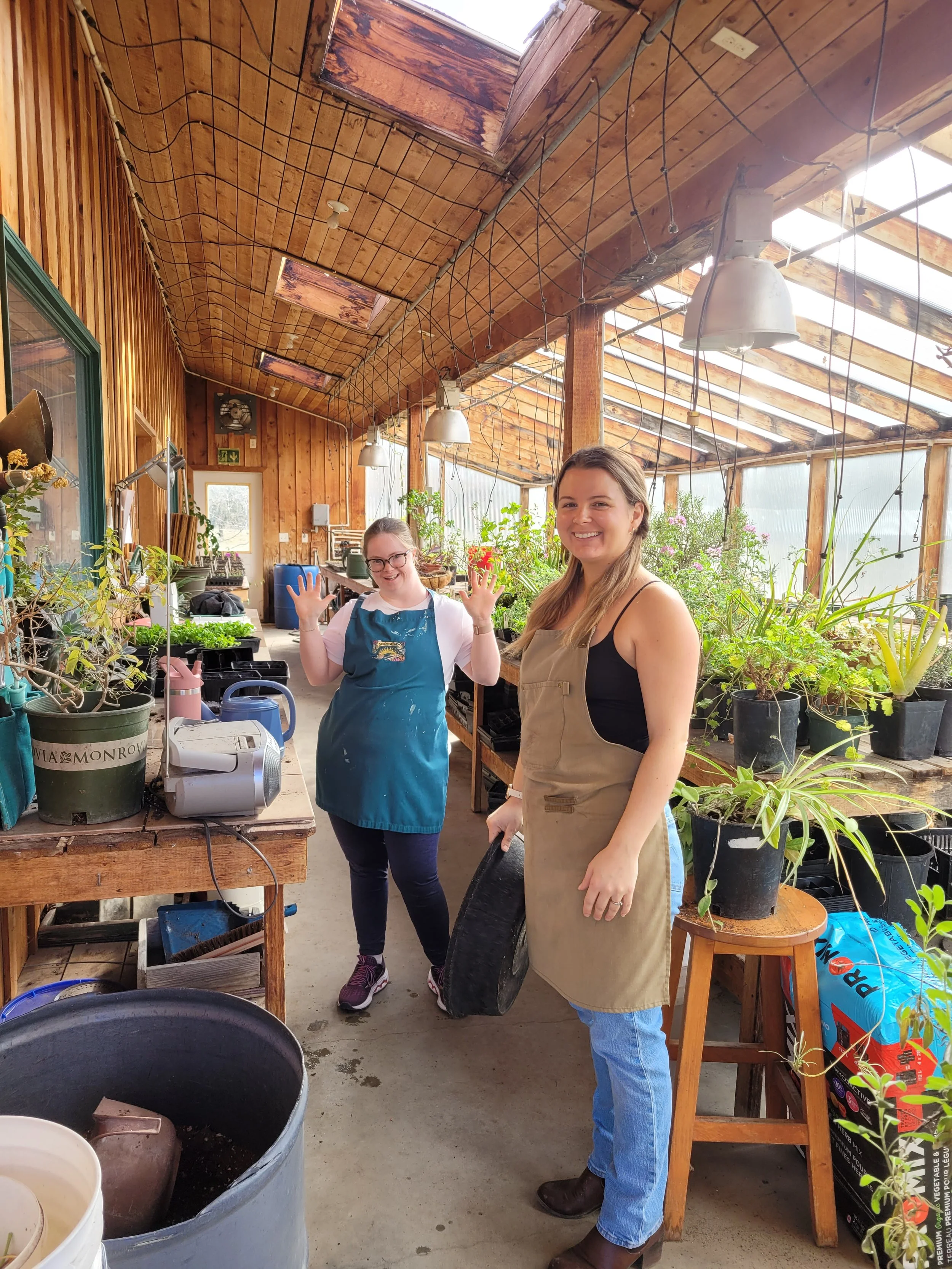 Two women inside a wooden greenhouse or garden shed with plants, tools, and gardening supplies, smiling at the camera.