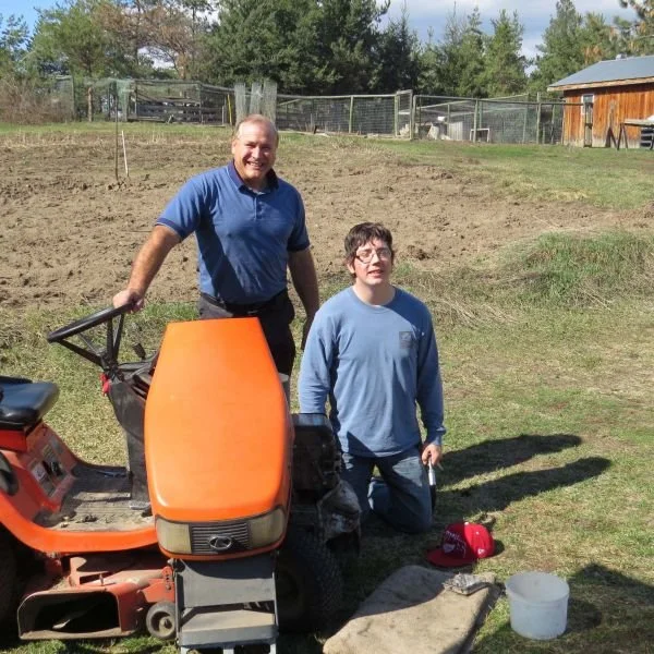 A man and a boy are outdoors on a grassy area with a dirt patch behind them. The man is standing and smiling, holding onto a small orange tractor or ride-on lawn mower. The boy is kneeling on one knee, wearing glasses and a blue shirt, holding a smal