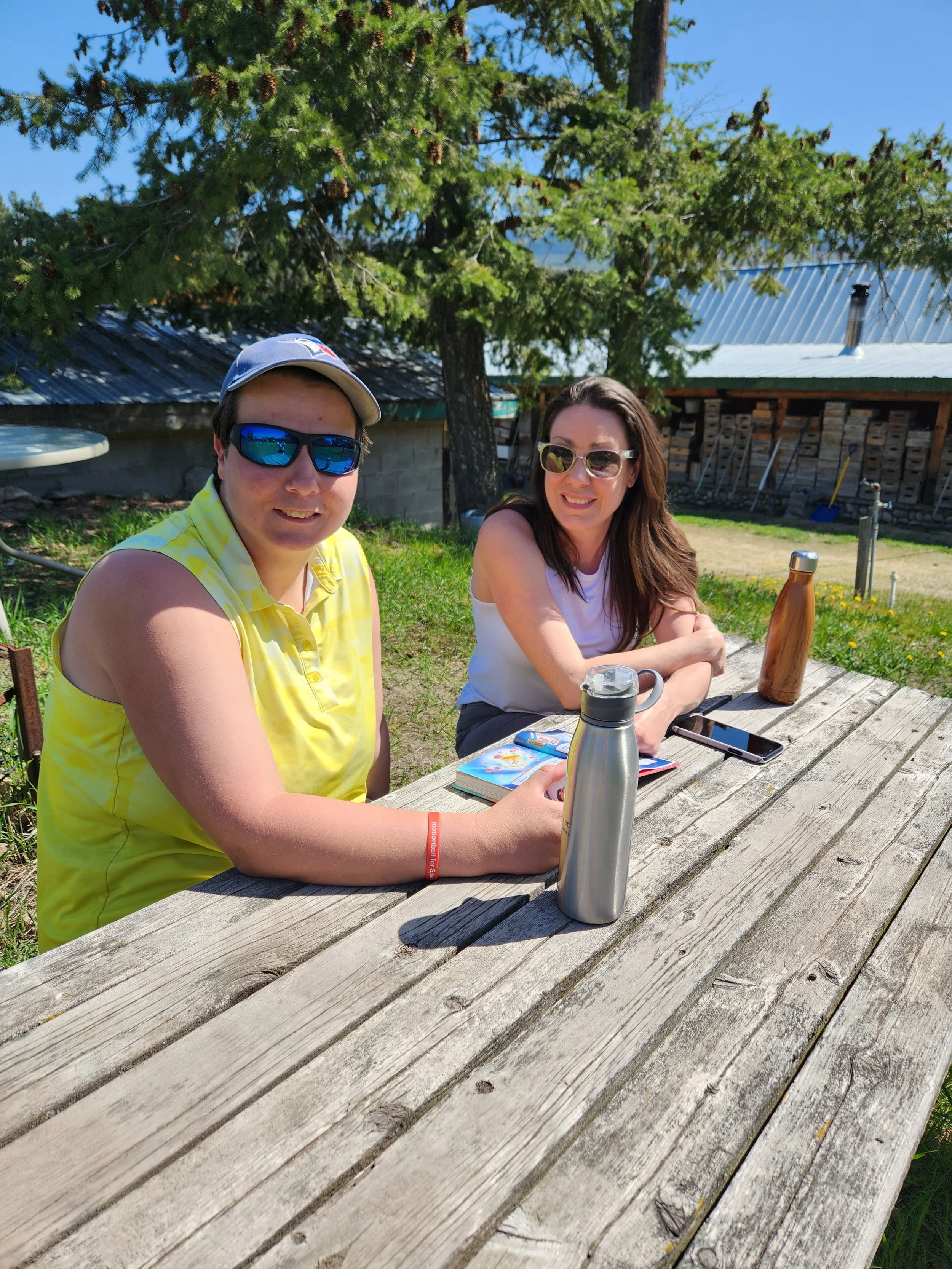 Two women sitting at a wooden picnic table outdoors on a sunny day, with water bottles and a book in front of them, and a tree and rustic building in the background.