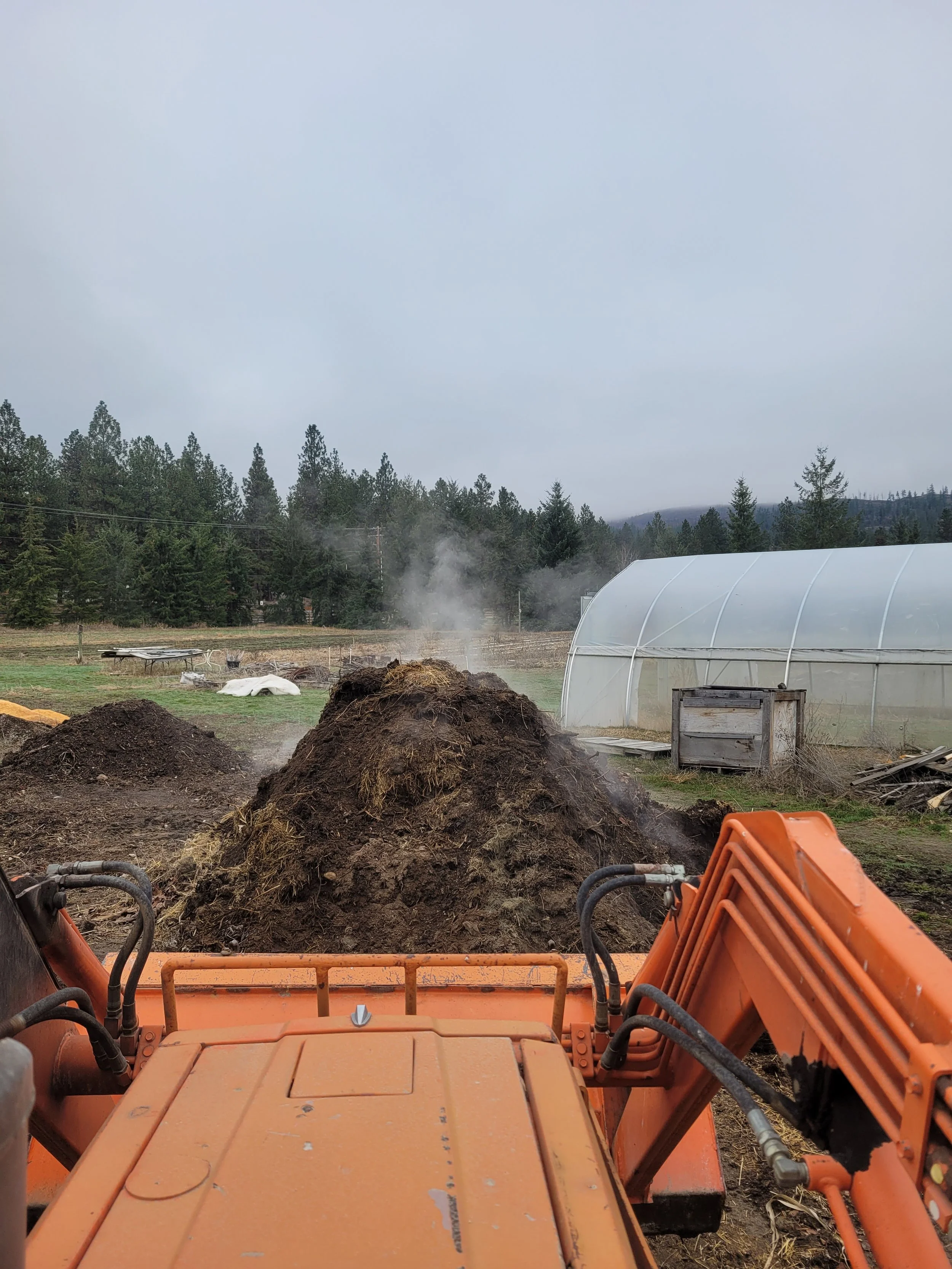 Agricultural scene with tractor in foreground, large compost pile, greenhouse, and forested hills in the background.