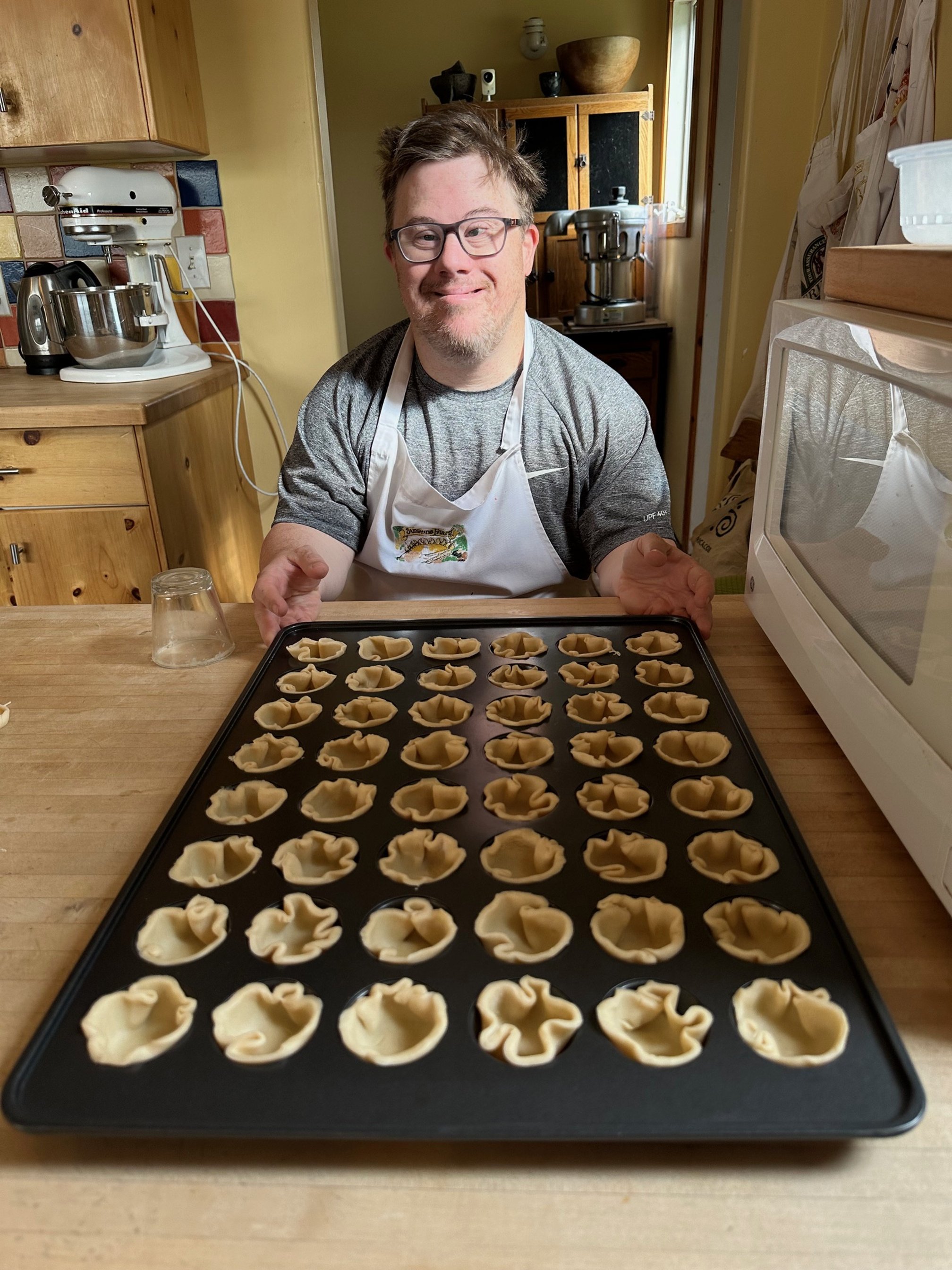 A man in glasses and a gray t-shirt, wearing an apron, sitting at a kitchen counter with a tray of uncooked tortellini pasta in front of him, smiling at the camera.