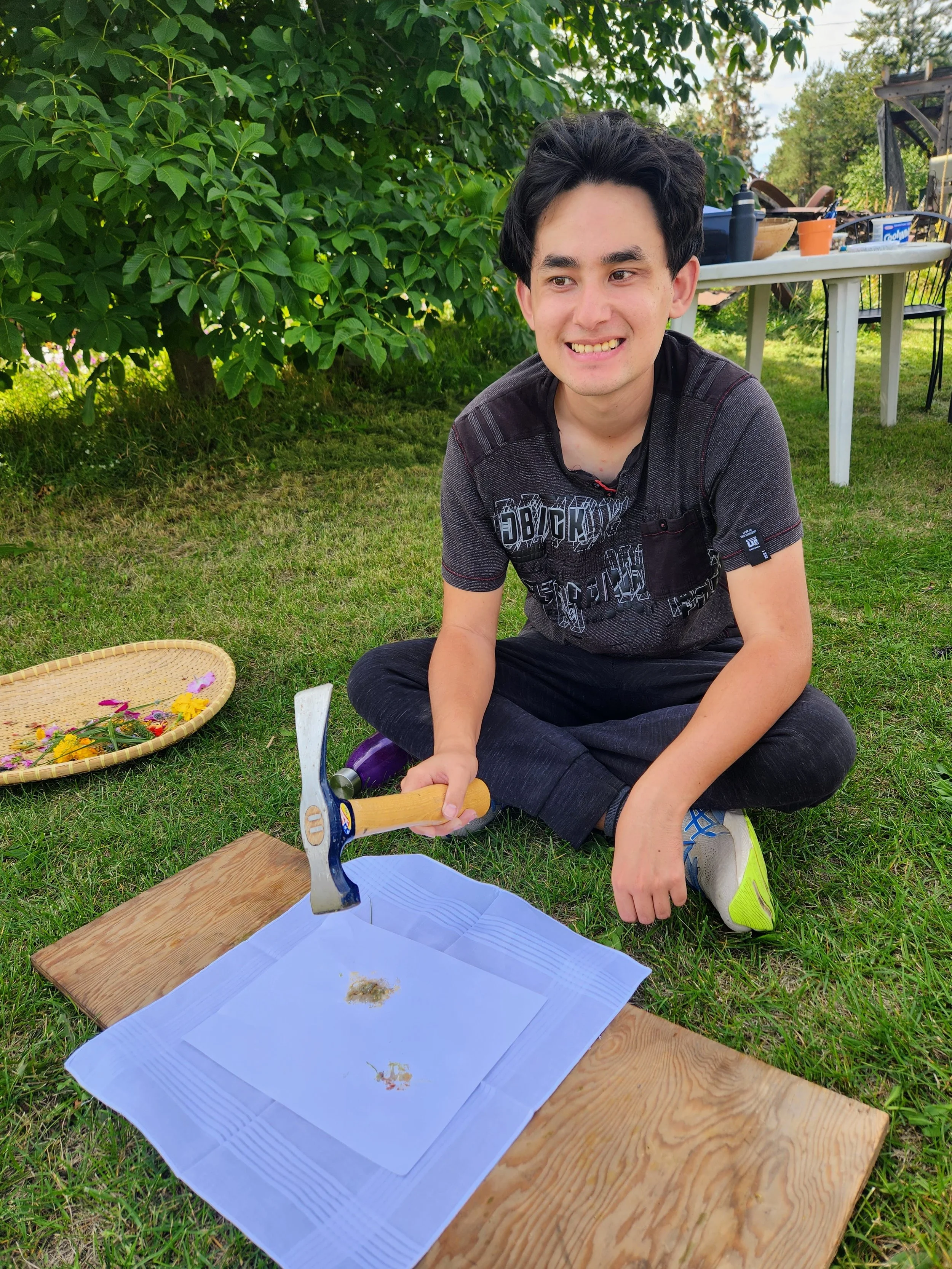A young man with black hair sits on the grass, smiling, holding an axe over a piece of wood, in a garden setting. There's a basket with flowers nearby and a table with supplies in the background.